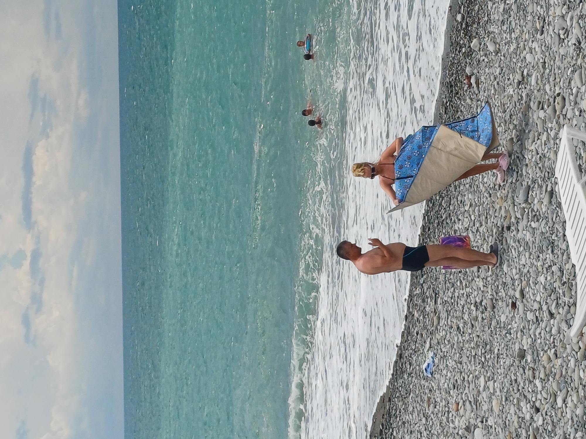 The image depicts a pebble beach in Sochi, Russia, with a wide expanse of turquoise water under a cloudy sky. In the foreground, a man in black swim trunks stands near the water's edge, holding a cigarette. To his right, a woman in a bikini is holding a patterned beach blanket, seemingly preparing to lay it down. The sea is active with moderate waves, and several people, including children, are visible swimming and playing in the water in the mid-ground. The beach is covered in smooth grey and white pebbles. A white lounge chair is partially visible in the bottom left corner of the frame.