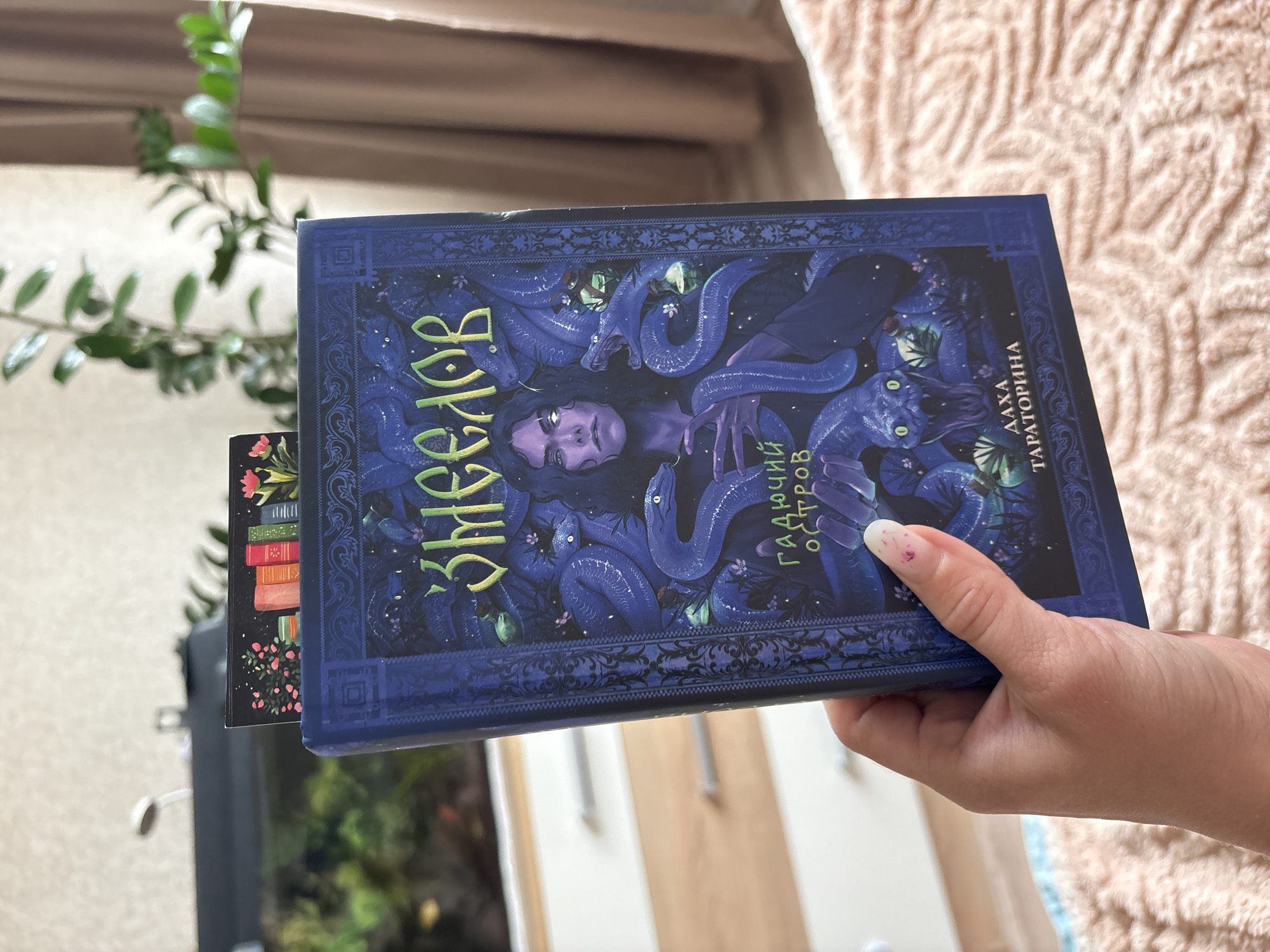 A hand holds an open book with a dark blue cover featuring an illustration of a person surrounded by snakes. The book is oriented vertically, with the spine facing the viewer. The cover art depicts a pale-skinned person with dark hair and glowing white eyes, their face turned slightly to the left. Various blue snakes are coiled around the person, some with green eyes. The title "Змеелов" is written in yellow, stylized Cyrillic script across the top of the cover. Below this, in green Cyrillic, are the words "ГАДЮЧИЙ ОСТРОВ". At the bottom of the cover, the author's name "ДАХА ТАРАТОРИНА" is also in green Cyrillic. A bookmark with an illustration of a stack of colorful books and pink flowers is partially visible sticking out from the top of the book. The background shows a blurred interior scene with a shelf or dresser unit on the left and a plant and beige curtains on the right. The floor appears to be covered with a textured, light-colored fabric, possibly a blanket or rug. The image is not taken in Noviy, Russia; there are no recognizable landmarks or indicators of this specific location.