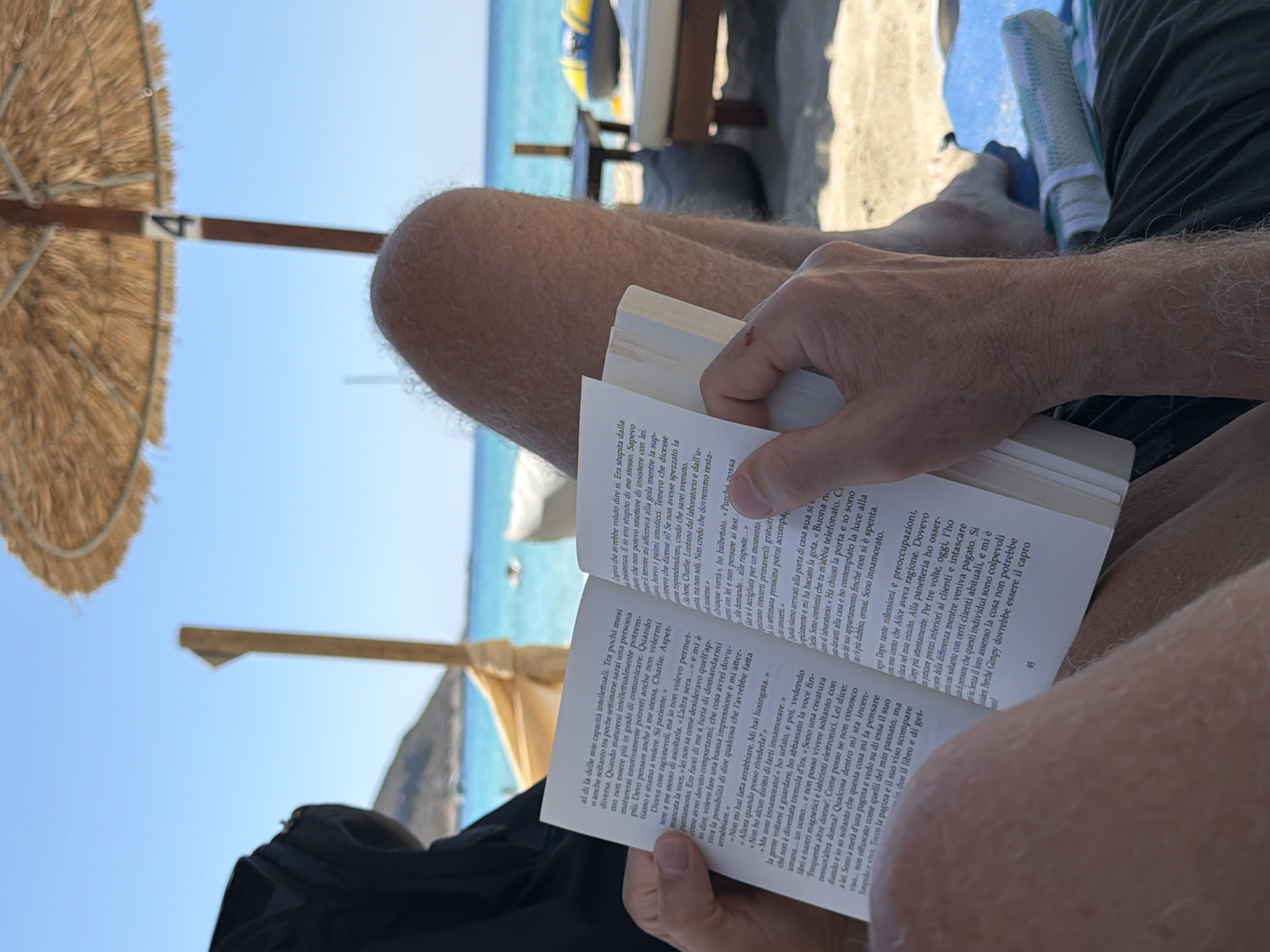 A person is sitting on a beach, reading a book, with their legs visible in the foreground. The person is wearing dark shorts and has hairy legs. The book is open and being held by the person's hands. In the background, there is a straw umbrella providing shade, a beach chair, and the turquoise sea. The location is a beach in Greece.
