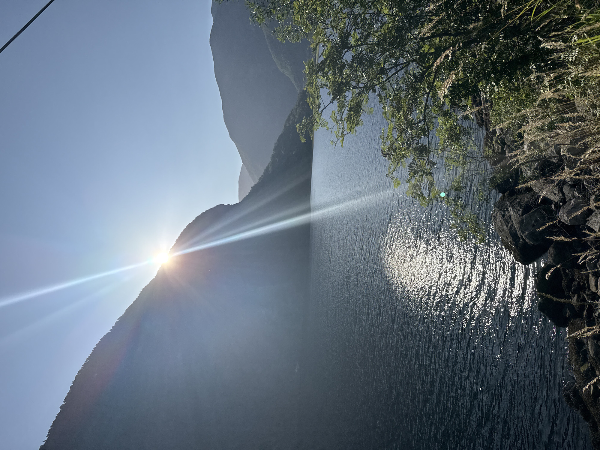 A wide shot captures a serene natural landscape under a bright, clear sky. The sun is positioned high, just peeking over the crest of a massive, tree-covered mountain that dominates the left side of the frame. Its rays dramatically fan out, creating a brilliant lens flare that streams across the image.
Below the mountain lies a vast, dark body of water, its surface rippled by a gentle breeze. The sun's reflection shimmers on the water, creating a sparkling path of light that stretches towards the foreground. In the distance, more undulating mountains, shrouded in a hazy blue, form a picturesque backdrop.
The right side of the image features a rocky shoreline. Overgrown with lush green foliage and tall, dry grasses, this area adds a touch of wildness to the scene. Several large, dark rocks are scattered along the edge of the water. The overall impression is one of peaceful, untouched nature. The scene is consistent with the natural beauty found in Lærdalsøyri, Norway, known for its dramatic fjords and mountainous terrain.