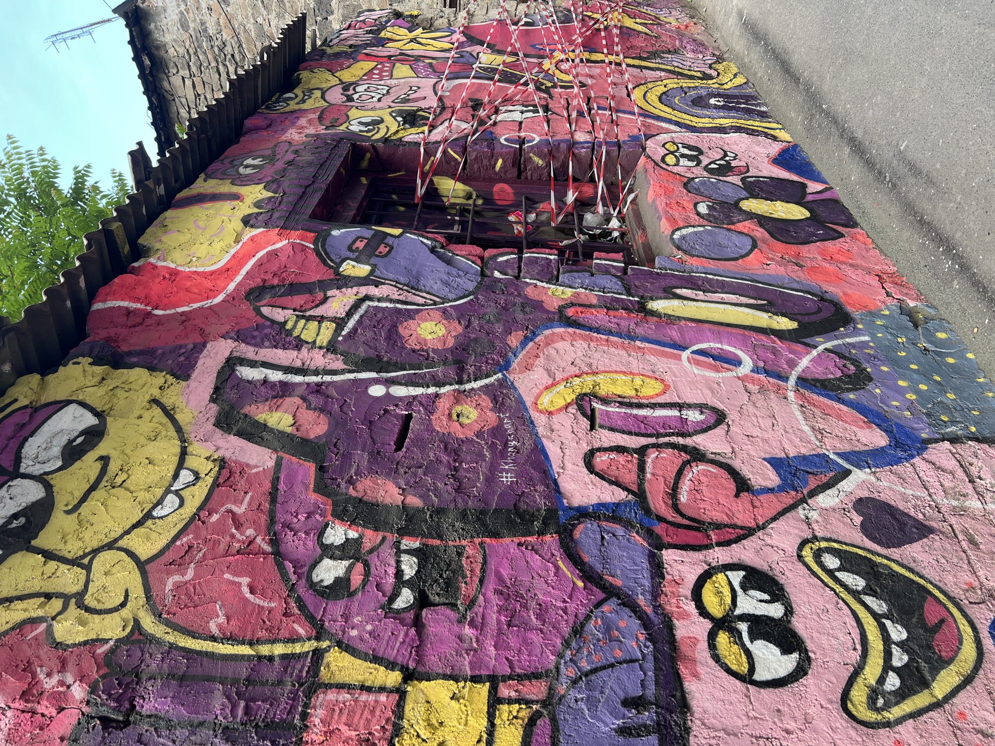 A low-angle, full-shot photograph showcases a weathered, graffiti-covered wall, likely part of a building, in Yerevan, Armenia. The wall's surface is deeply textured with cracks and uneven sections, coated in a vibrant array of colors, including yellow, purple, pink, and red. The graffiti itself features cartoonish figures and abstract patterns. A hole exists in the wall, and safety tape covers the hole. The building's roof is visible at the top, constructed from stone and timber. The environment consists of the sky and a concrete surface.
