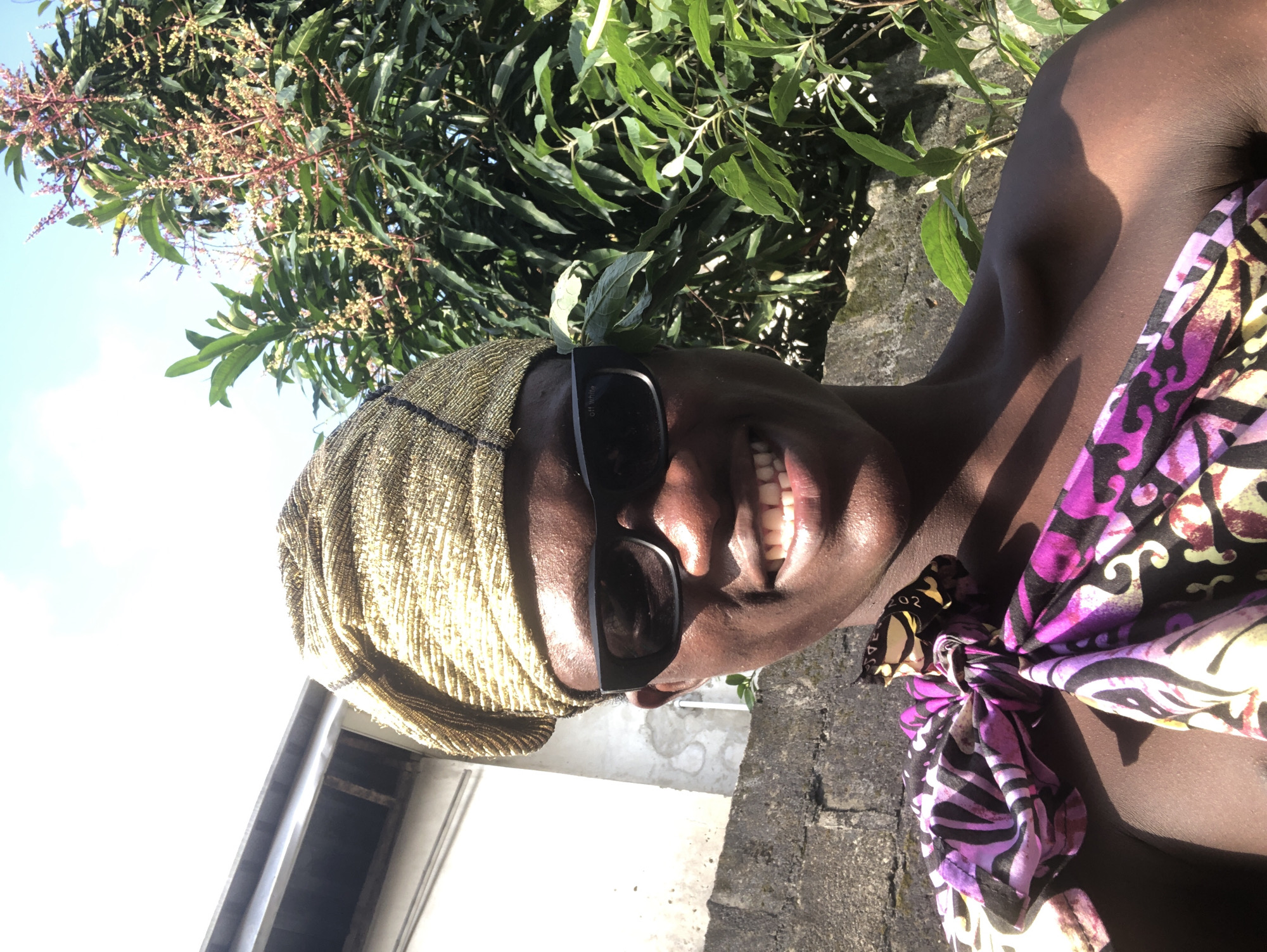 A person, likely male, is seen from the chest up, taking a selfie outdoors in what appears to be a residential area. The individual is wearing a golden headwrap and black sunglasses. A brightly colored fabric, predominantly purple, pink, and black, is draped over the person's shoulder. The background reveals a stone wall, a building with a roof, and lush green foliage from a tree, possibly a mango tree with developing fruit. The sky above is bright and clear, suggesting a sunny day. The location is identified as Ado, Nigeria.