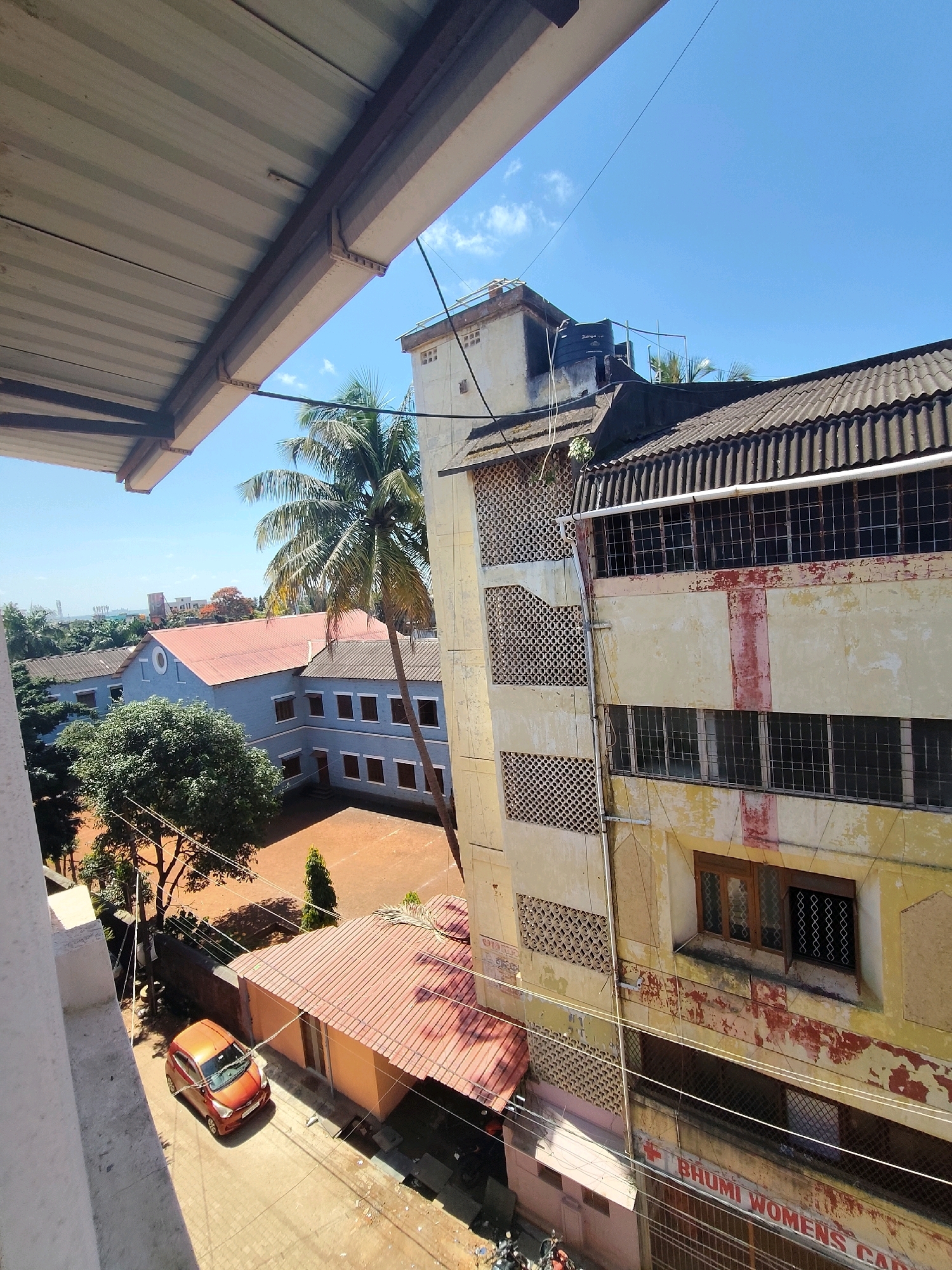 An aerial view captures a bright, sunny day in the city of Belagavi, India. The foreground features a corrugated metal overhang, casting a shadow. Below, a street with a paved surface is visible, with an orange car parked on the left. To the right of the street, a building with a red-tiled roof and an orange-colored facade is situated.
Further back, a large, light blue building with a red roof dominates the middle ground. This building has multiple windows and is surrounded by trees, including a prominent palm tree. Adjacent to it, a taller, multi-story building with a weathered yellow facade and dark-tiled roof stands out. This building features numerous windows, some with bars, and a distinctive pattern of circular perforations on its exterior. A faded red stripe runs vertically down its facade.
In the background, more buildings and greenery are visible, with a clear blue sky overhead. The scene suggests an urban environment with a mix of residential and commercial structures, typical of a city in India. The overall impression is one of a developed area under clear weather conditions.