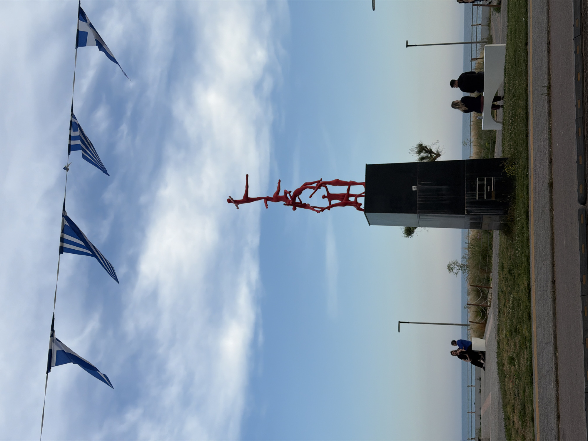 The image displays a coastal scene in Alexandroupolis, Greece, under a partly cloudy sky. The main subject is a prominent modern sculpture centrally located, composed of numerous abstract, red human figures that appear to be intertwined or stacked, forming an upward-stretching, complex shape. This sculpture rests on a dark, rectangular pedestal.
To the upper left, several Greek flags are strung across the sky. These triangular pennants feature the characteristic blue and white stripes with a white cross on a blue canton. The sky transitions from a lighter blue near the horizon to a deeper blue above, dotted with scattered white clouds, suggesting a pleasant, partly overcast day.
On the right side of the image, a paved promenade runs along the coastline. Adjacent to the promenade, there's a strip of green grass and some sparse vegetation. In the background, the calm, light blue sea extends to the horizon.
Several people are visible on the promenade. In the mid-ground, a man and a woman are seated on a modern white bench, facing the sea. Further along the promenade, another two individuals can be seen walking or standing near the water's edge. The scene depicts a public waterfront area, suggesting a leisurely atmosphere. No visible text is present on the sculpture or elsewhere in the image.