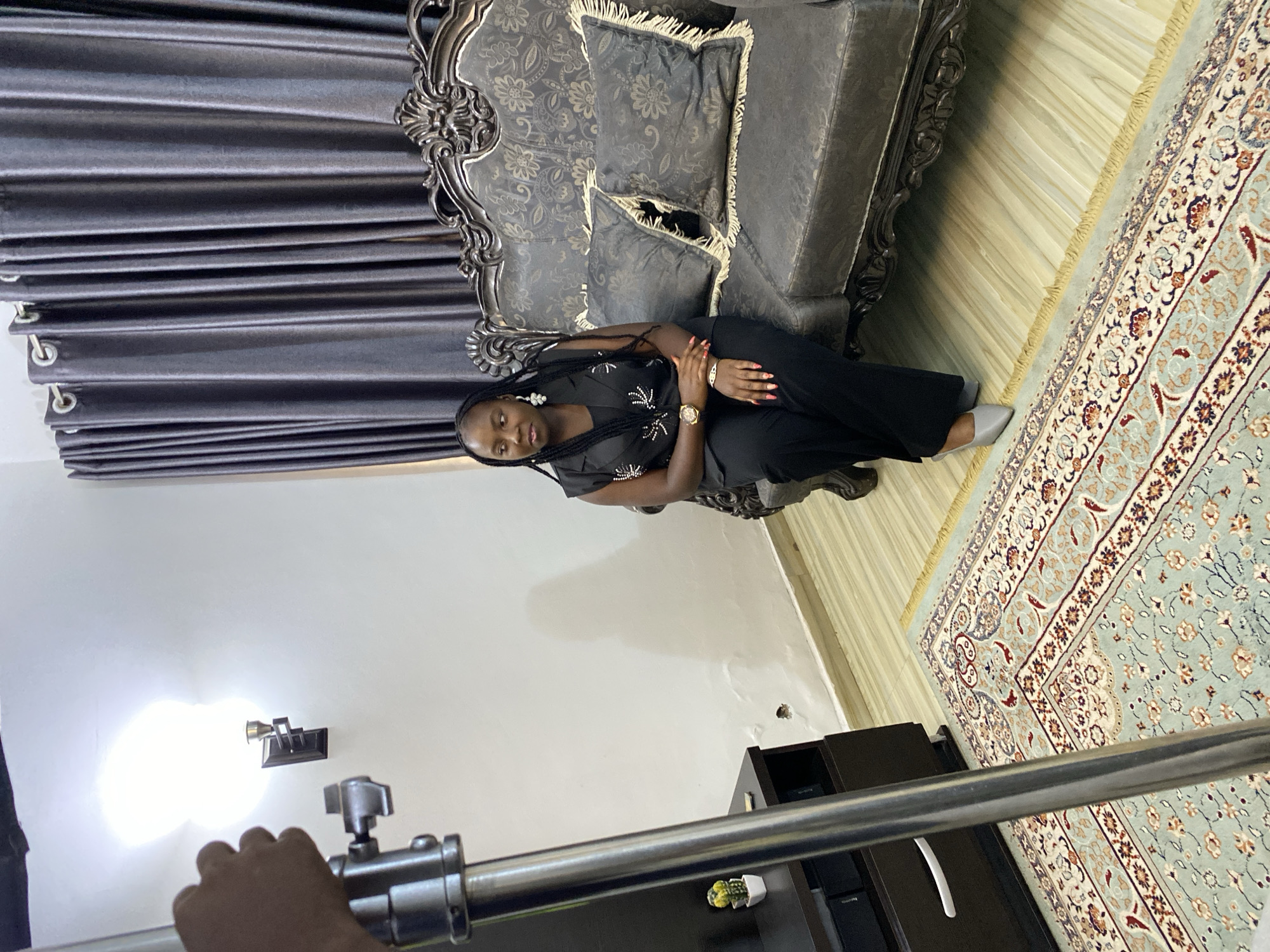 This media file captures an indoor scene, likely a living room or a professional studio setting, with a focus on a woman seated on an ornate sofa. The image, oriented sideways, shows a Black woman with braided hair, wearing a black top with white embellishments and black pants. She has a watch on her left wrist and red nail polish, and her expression is neutral as she looks slightly downwards.
The sofa is dark-colored with intricate carved details on its wooden frame and patterned fabric cushions featuring fringe. Behind the sofa are dark grey, pleated curtains. The floor beneath the sofa is a light-colored wood or tile, partially covered by a light blue/green patterned rug with floral and geometric designs and fringed edges.
In the foreground, part of a metallic stand or boom arm, likely belonging to lighting or camera equipment, is visible, held by a hand. A bright beam of light illuminates a white wall, emanating from a small fixture on the wall, suggesting a controlled lighting setup. A dark cabinet or shelf unit is partially seen in the lower right, holding a small decorative potted plant. The presence of professional equipment indicates a staged event, possibly a photoshoot or video recording, in an indoor environment, potentially in Jos, Nigeria. No visible text is present in the image.