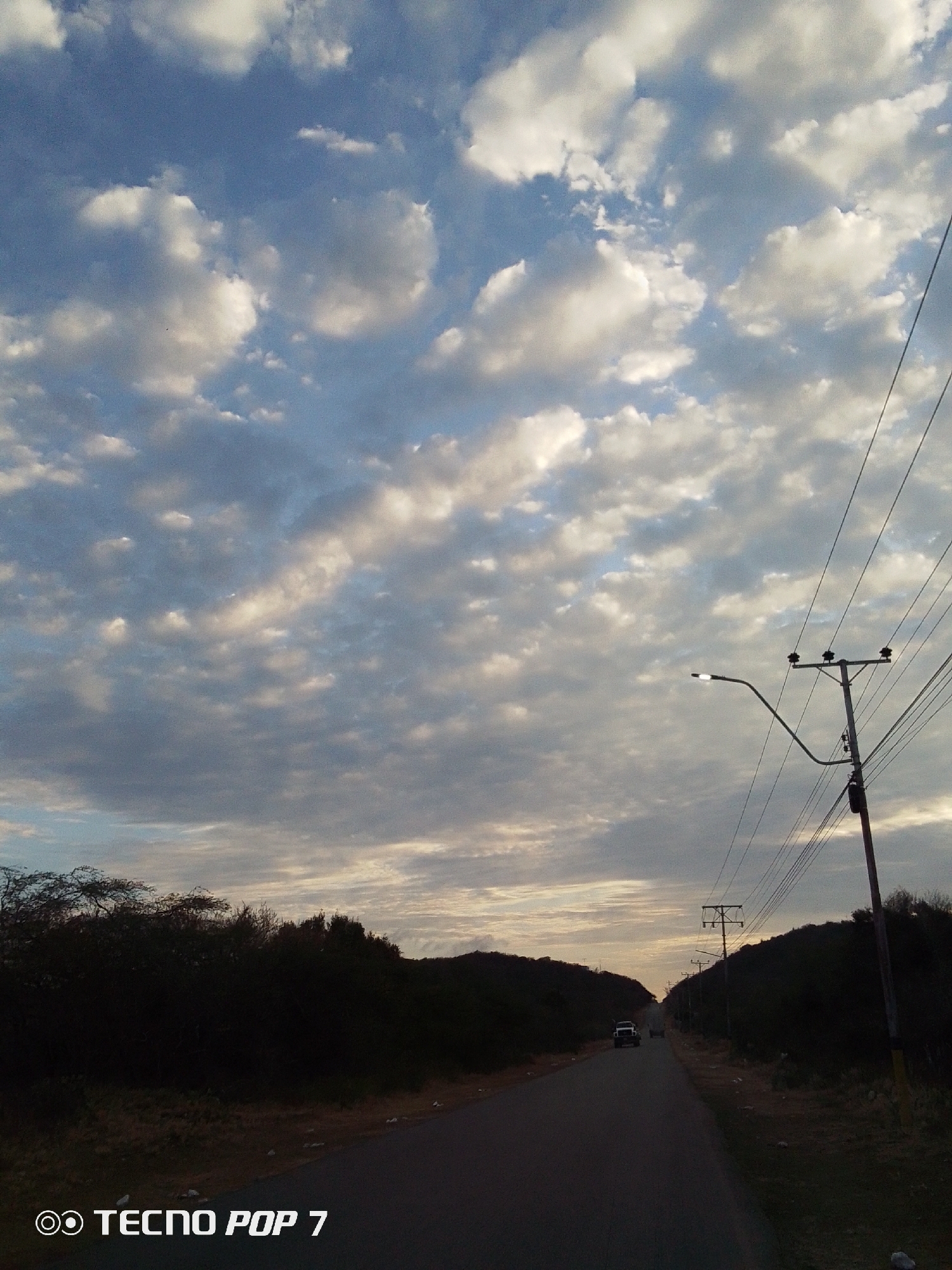 The image depicts a road leading into the distance, flanked by dark vegetation on both sides. Above, a sky filled with clouds is visible, with hints of sunlight breaking through. Utility poles and power lines run along the side of the road. A vehicle can be seen in the distance. The location is implied to be Altagracia, Venezuela.