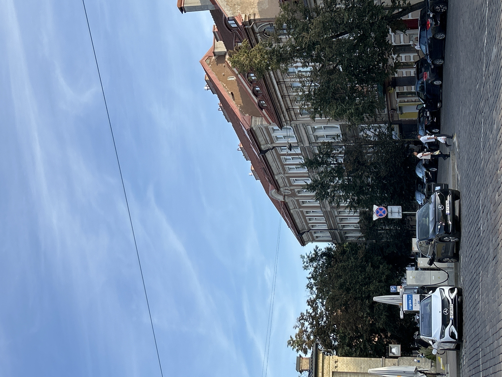 The image depicts a street scene in Vilnius, Lithuania, likely in the Old Town, given the architecture. The foreground shows a cobblestone street with several parked cars, including two electric vehicles being charged at a charging station. One is a white Mercedes-Benz, and the other is a black Mercedes-Benz. A few people are walking on the sidewalk, some carrying shopping bags.
In the background, there are several multi-story buildings with traditional European architecture, characterized by red-tiled roofs and ornate facades. Trees line the street, providing shade. The sky is blue with wispy clouds, and a power line stretches across the upper portion of the frame. A street sign with a blue "P" indicating parking is visible, as is a circular red and blue sign. The overall impression is of a peaceful urban environment on a clear day.