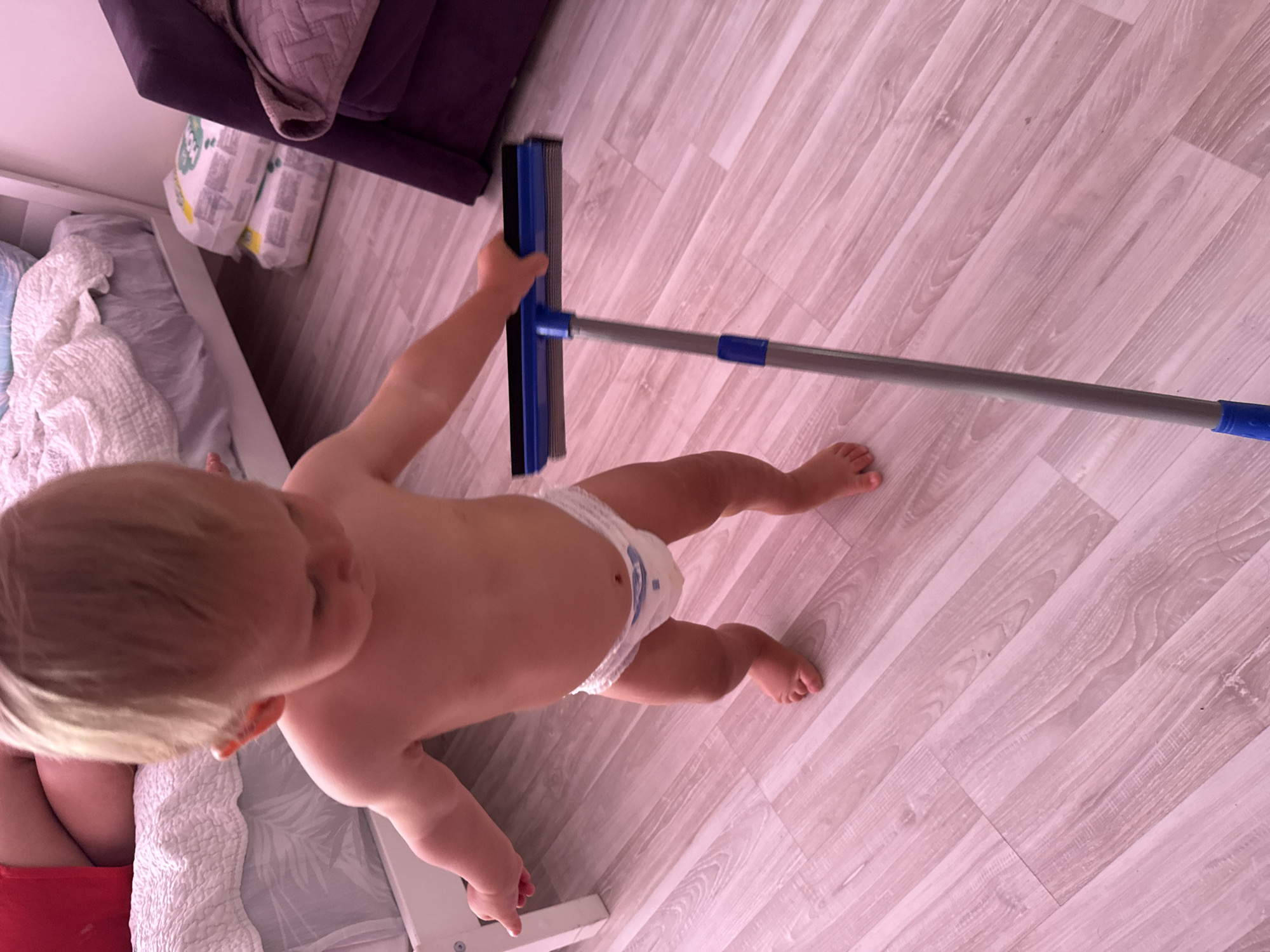 A toddler, wearing only a diaper, stands on a light-colored, wood-plank floor and holds a blue and gray squeegee. The toddler is facing away from the camera, looking down at the squeegee. The toddler has blond hair and is facing towards the right side of the frame. To the left of the toddler, a bed with white bedding is partially visible. In the background, on the right side of the image, a purple armchair and two stacked white packages of diapers are visible. The lighting in the image is dim and appears to be pinkish, possibly due to ambient lighting. The scene does not contain any recognizable landmarks or elements that would specifically place it in Sochi, Russia.
