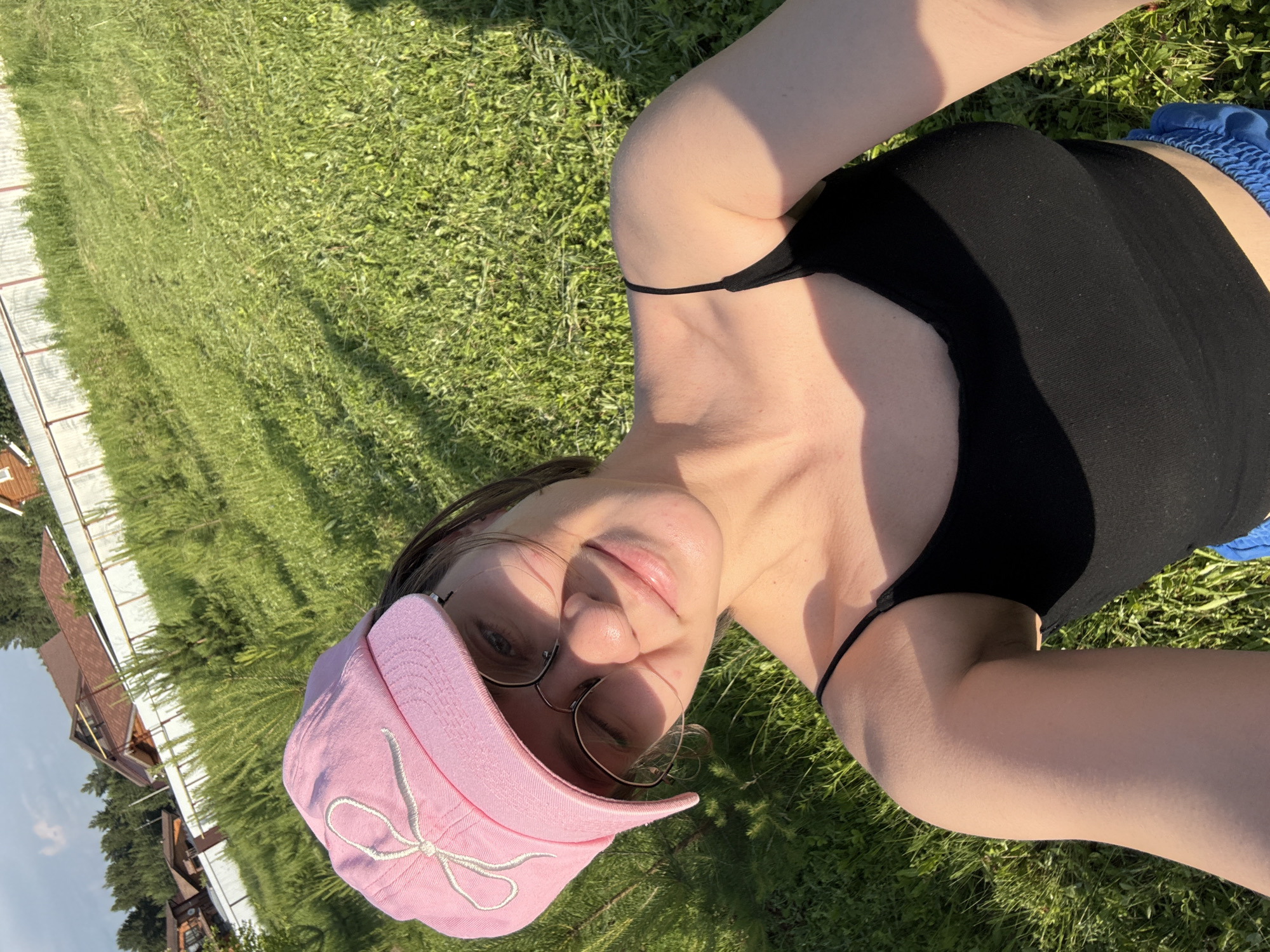 A young woman with brown hair and glasses is taking a selfie outdoors in a grassy area. She is wearing a pink baseball cap with a white bow embroidered on the front, a black tank top, and blue shorts. The background shows a residential area with houses and a white fence, possibly in the outskirts of Dokshitsa, Russia. The lighting suggests it is daytime, with sunlight casting shadows on the grass.