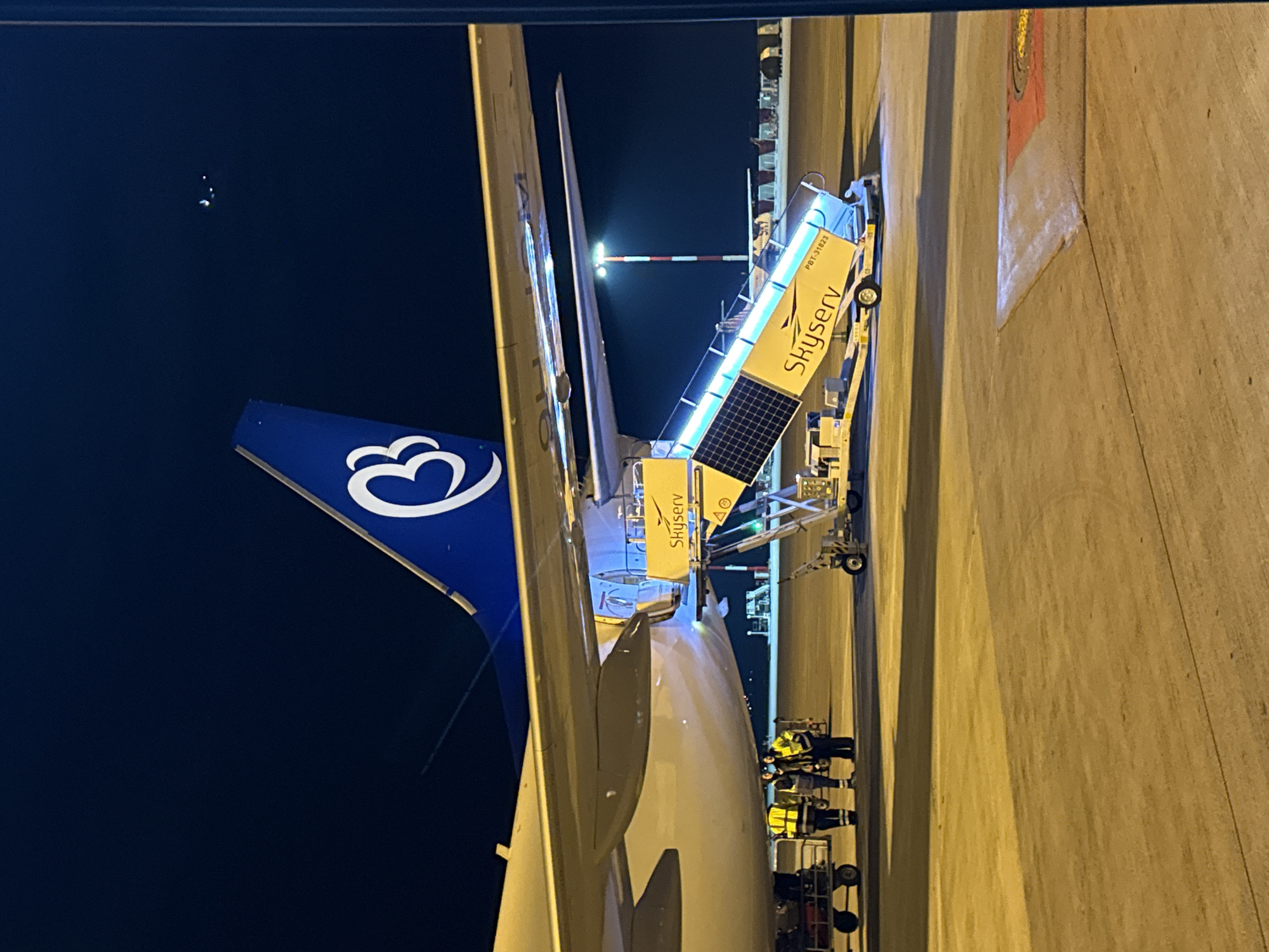 The image depicts an nighttime scene on the tarmac of Athens International Airport "Eleftherios Venizelos" in Paiania Municipal Unit, Greece. A large passenger aircraft, identifiable by its blue tail featuring a white stylized 'B' or wing-like logo (consistent with Aegean Airlines/Olympic Air), is parked on the illuminated concrete apron. A mobile passenger stair, branded "Skyserv" and labeled "PBT-31623," is positioned against an open aircraft door, illuminated by blue LED lights and equipped with visible solar panels. To the left of the stairs, approximately three ground crew personnel, wearing high-visibility yellow jackets and face masks, are gathered near the aircraft's fuselage, adjacent to two baggage carts or equipment trolleys. The background includes a tall, red and white striped airport light mast with an active light source, and distant airport infrastructure under a dark night sky.