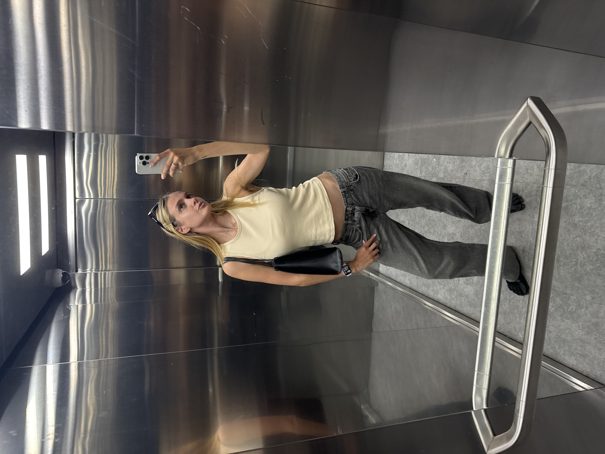 A person takes a photo in an elevator. The subject is a woman with long, blonde hair, fair skin, wearing a tan tank top, grey jeans, and black slides. She is holding a white phone in her right hand to take a selfie. She has a black purse hanging on her left shoulder. The elevator appears to be made of metal with fluorescent lights on the side. A silver handrail is visible on the right side. The location is Unknown, Unknown.