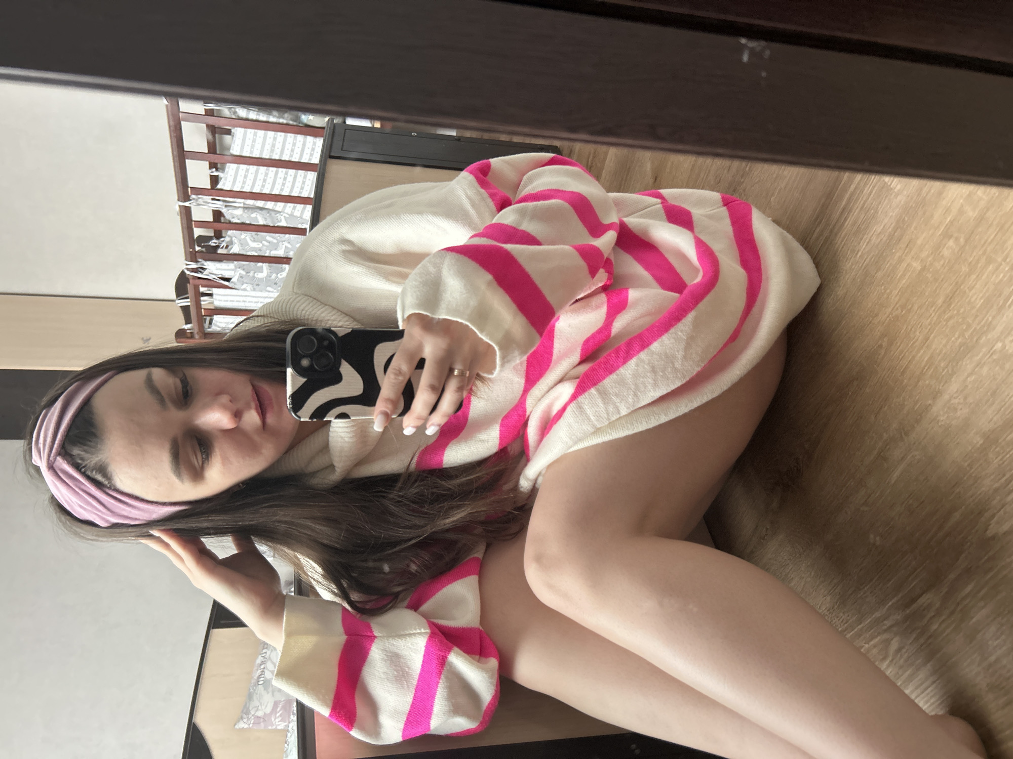 A young woman is captured in a mirror selfie, lying on a light wooden floor in what appears to be an indoor setting in Chaikovsky, Russia. She is positioned horizontally, looking upwards at her phone, which she holds in her right hand. Her eyes are partially closed, conveying a relaxed or contemplative mood.
She is wearing an oversized, cream or off-white knit sweater featuring broad, bright pink horizontal stripes, especially visible on the sleeves and main body. A dusty rose or light purple fabric headband adorns her head, holding back her dark hair. A ring is visible on the ring finger of her right hand. Her phone has a distinctive black and white wavy-patterned case. Her legs are bare and bent.
The background, visible as a reflection in the mirror, reveals elements of a domestic space. Most notably, a wooden crib with patterned bedding (grey fabric with white stars and animal figures) is visible, suggesting the room might be a nursery or a child's room. A plain, light-colored wall is also reflected. The top edge of the image shows the dark frame of the mirror. The lighting is soft and even, typical of an indoor daytime setting. There is no visible text in the image.