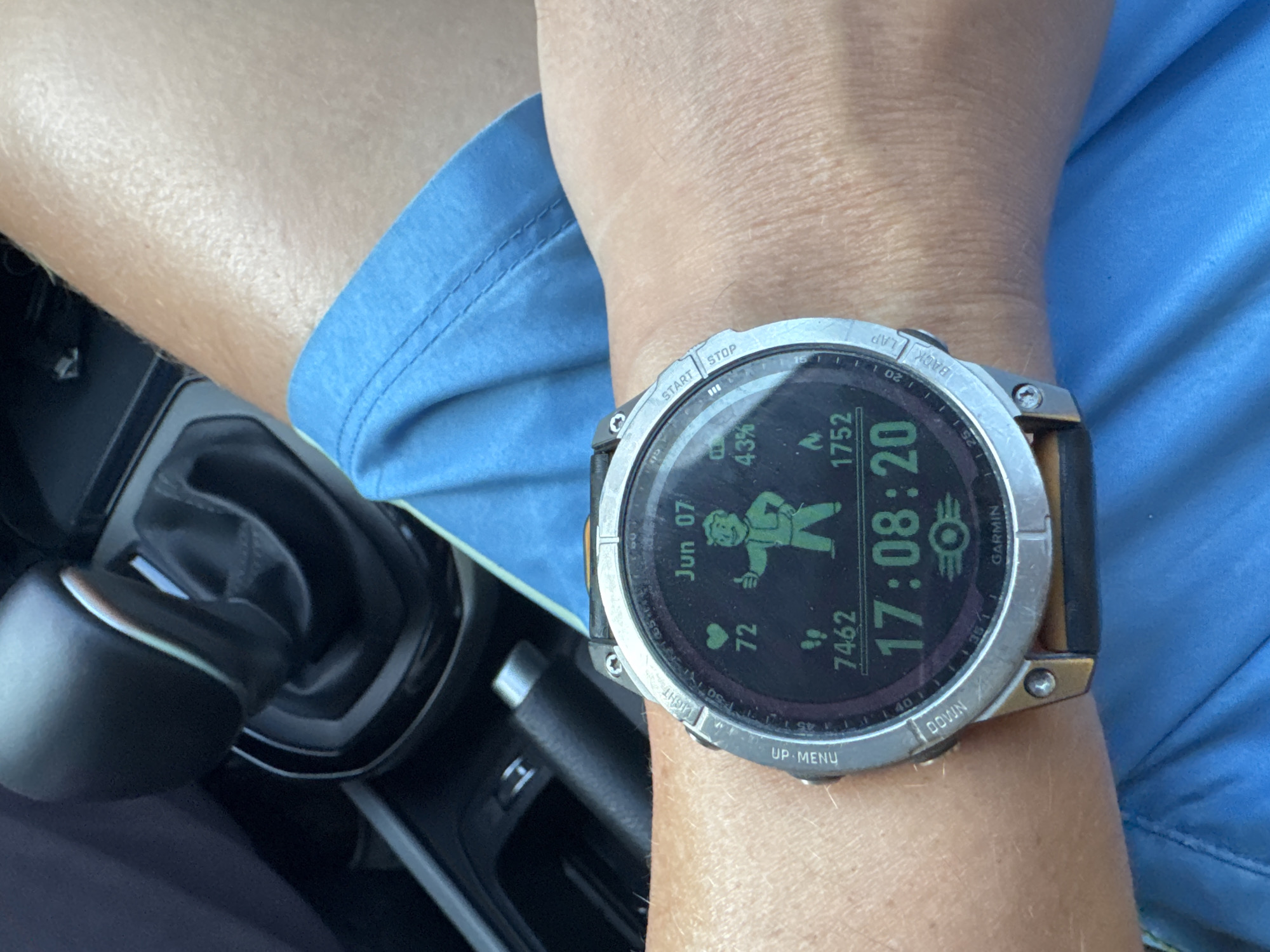 A fair-skinned forearm, likely male, is positioned in the foreground, wearing a Garmin smartwatch on the left wrist (relative to the viewer). The arm is partially covered by a light blue short-sleeved garment, displaying visible stitching. The smartwatch features a silver-toned bezel with engraved labels for buttons ("UP MENU," "LIGHT," "START STOP," "BACK LAP," "DOWN") and a dark strap. Its digital display, illuminated with green text on a dark background, shows "Jun 07" at the top, "17:08:20" (5:08 PM and 20 seconds) prominently in the center, and the Garmin logo at the bottom. Other data points include a heart rate of "72," a battery indicator at "43%," a step count of "7462," and calories burned at "1752." A green depiction of Fallout's Vault Boy character, giving a thumbs-up gesture, is central to the display.
In the background, the interior of a vehicle is visible, featuring a black automatic transmission gear selector with a silver-colored accent. To the left of the gear selector, a black, ribbed manual parking brake handle is present. The surrounding interior surfaces appear to be dark-colored plastic or upholstery. The scene is located in Nha Trang Ward, Vietnam.