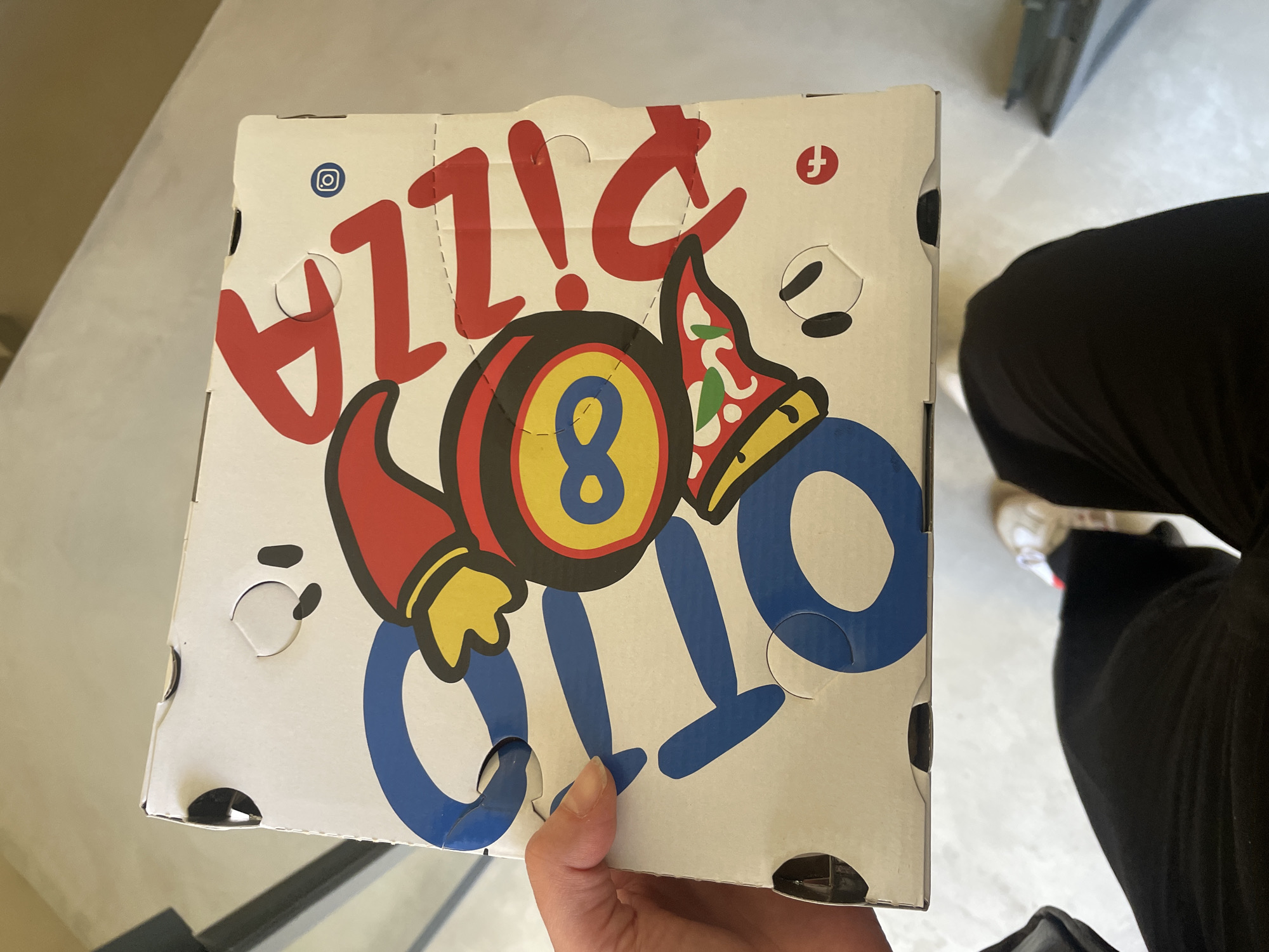 A person is holding a pizza box in their hand. The box is white with red and blue lettering that spells out "PIZZA! OTTO". The "O" in "OTTO" is an eight ball with a slice of pizza wearing a party hat on top of it. There are Instagram and Facebook logos on the box. The person is wearing black pants and white and red shoes. The scene is indoors. Based on the brand "OTTO", the location is likely Milan, Italy.