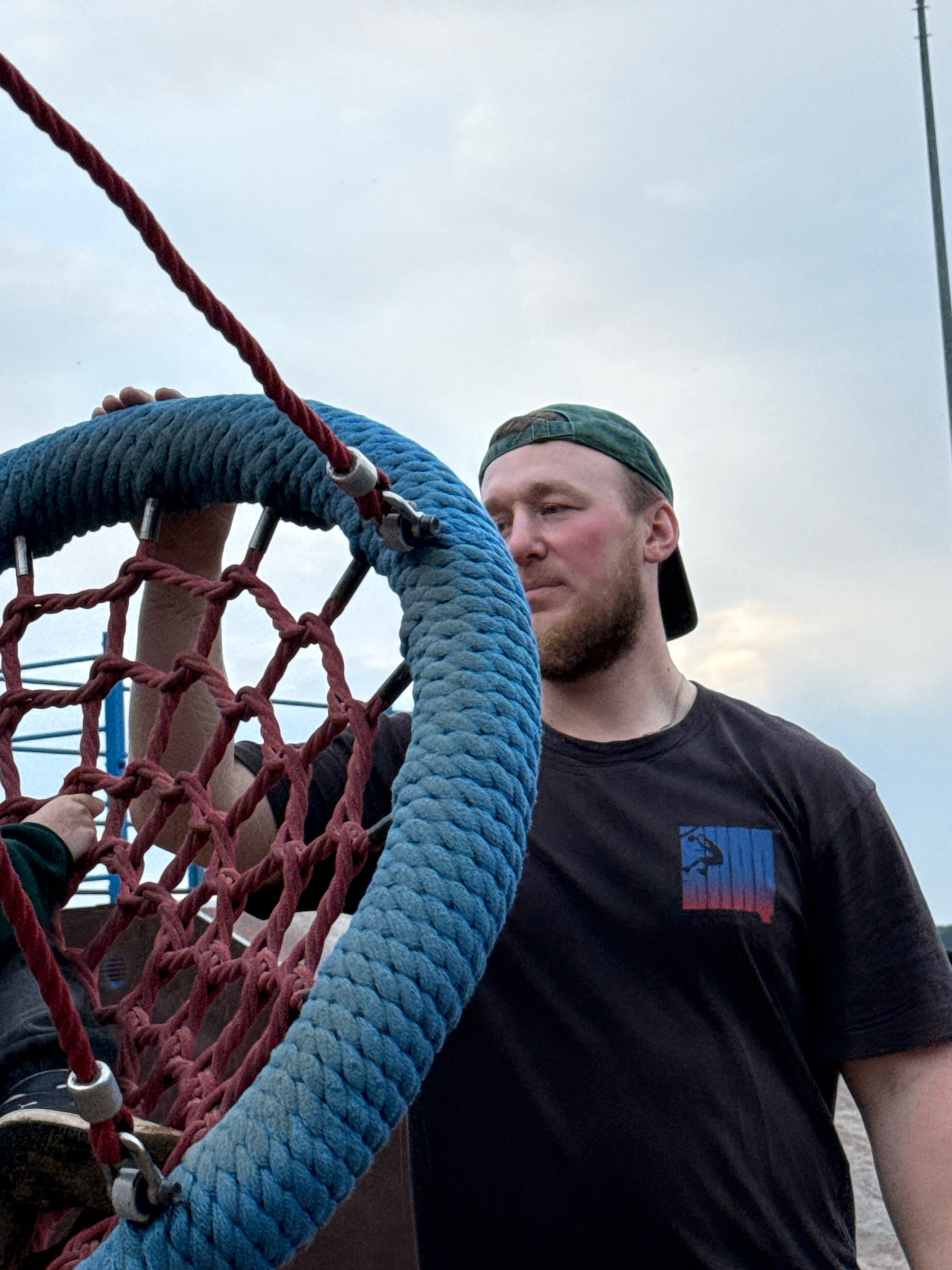A man wearing a black t-shirt and a green baseball cap is standing outdoors next to a large rope swing. The swing is made of thick blue rope forming the outer ring and a net of red rope for the seating area. The man is looking to the right, and his expression is neutral. His beard is short, and he has reddish hair. The t-shirt has a small graphic on the left side of the chest, depicting a silhouetted figure against a sunset. The background consists of a cloudy sky and a tall, thin structure, possibly a pole or antenna, on the right side. The ground appears to be sandy or gravelly. The scene is likely in Chaikovsky, Russia, given the context of the provided information.