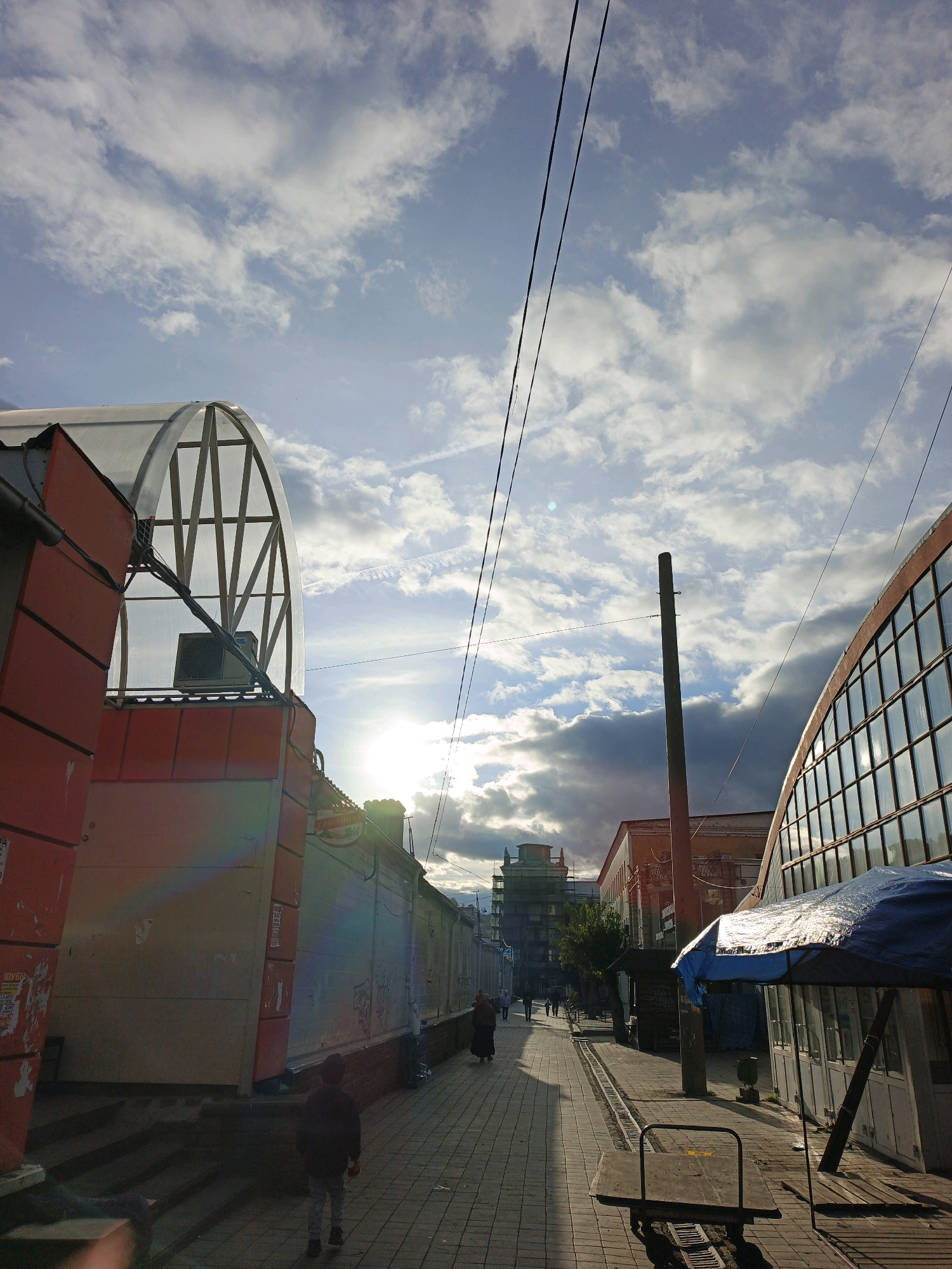 In Zlatoust, Russia, on a paved walkway, a person is walking away from the camera towards a street lined with buildings and market stalls. The sun is setting, casting long shadows and illuminating the clouds in the sky. The buildings on either side of the street appear to be commercial structures, with one on the right featuring a curved glass facade and a blue tarped market stall in front of it. Overhead power lines crisscross the sky. The overall scene suggests an outdoor market area in a city.