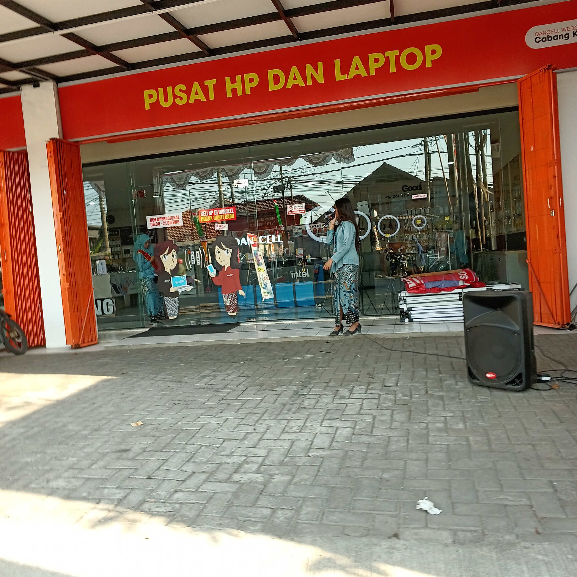 The image depicts the exterior of a retail store with a large glass storefront. A bright orange awning with yellow lettering that reads "PUSAT HP DAN LAPTOP" (meaning "Center for Phones and Laptops") is prominently displayed above the entrance. To the right, a smaller sign in white lettering on a black background reads "DANCELL WEDORO" and below it, "Cabang Ki".

Inside the store, visible through the glass, are several figures. Two cartoonish cutouts of women are positioned near the front. One is holding a smartphone and wearing a red shirt and a patterned skirt. The other is wearing a blue hijab and a patterned skirt. A real person, a woman wearing a light blue hijab and a patterned skirt, stands near the window, holding a microphone and speaking. She is facing towards the right side of the image.

In front of the store, on the paved ground, is a black speaker. Stacks of flat, white objects, possibly boxes, are placed to the left of the speaker. A portion of a motorcycle wheel and tire is visible on the far left of the image. The ground is paved with grey interlocking bricks. The overall impression is a street-level view of a commercial establishment, likely in a town or city. The mention of "WEDORO" on the sign indicates the specific location within Indonesia.