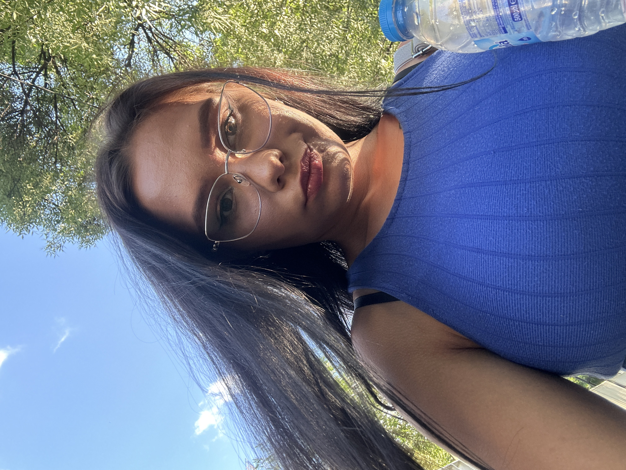 The image is a selfie taken outdoors, under a blue sky with some clouds, and a backdrop of green trees. The subject is a young woman with long, dark hair and glasses. She is wearing a blue ribbed tank top. To her right, a plastic bottle of water with a blue cap is visible. The lighting suggests it is daytime. The scene is not identifiable as being in Sofia, Bulgaria.