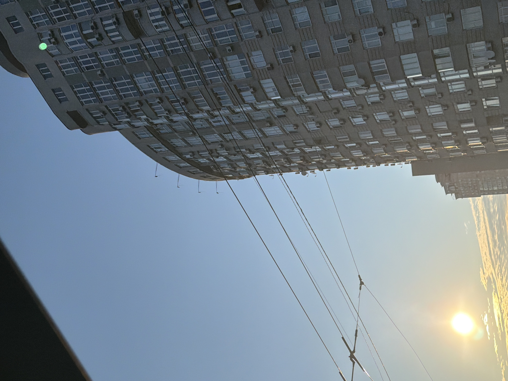 An angled, eye-level medium shot captures a modern, light gray, high-rise apartment building with a curved facade against a clear, pale blue sky. The building's exterior is detailed with numerous windows and air conditioning units. Several dark, overhead wires stretch across the frame from the left foreground, obscuring parts of the sky and the building. On the far left, a bright, white sun radiates a lens flare, illuminating a strip of soft, yellow clouds below. A smaller, dark building is partially visible to the left of the main apartment building. A small, bright green light is visible on the top corner of the apartment building.