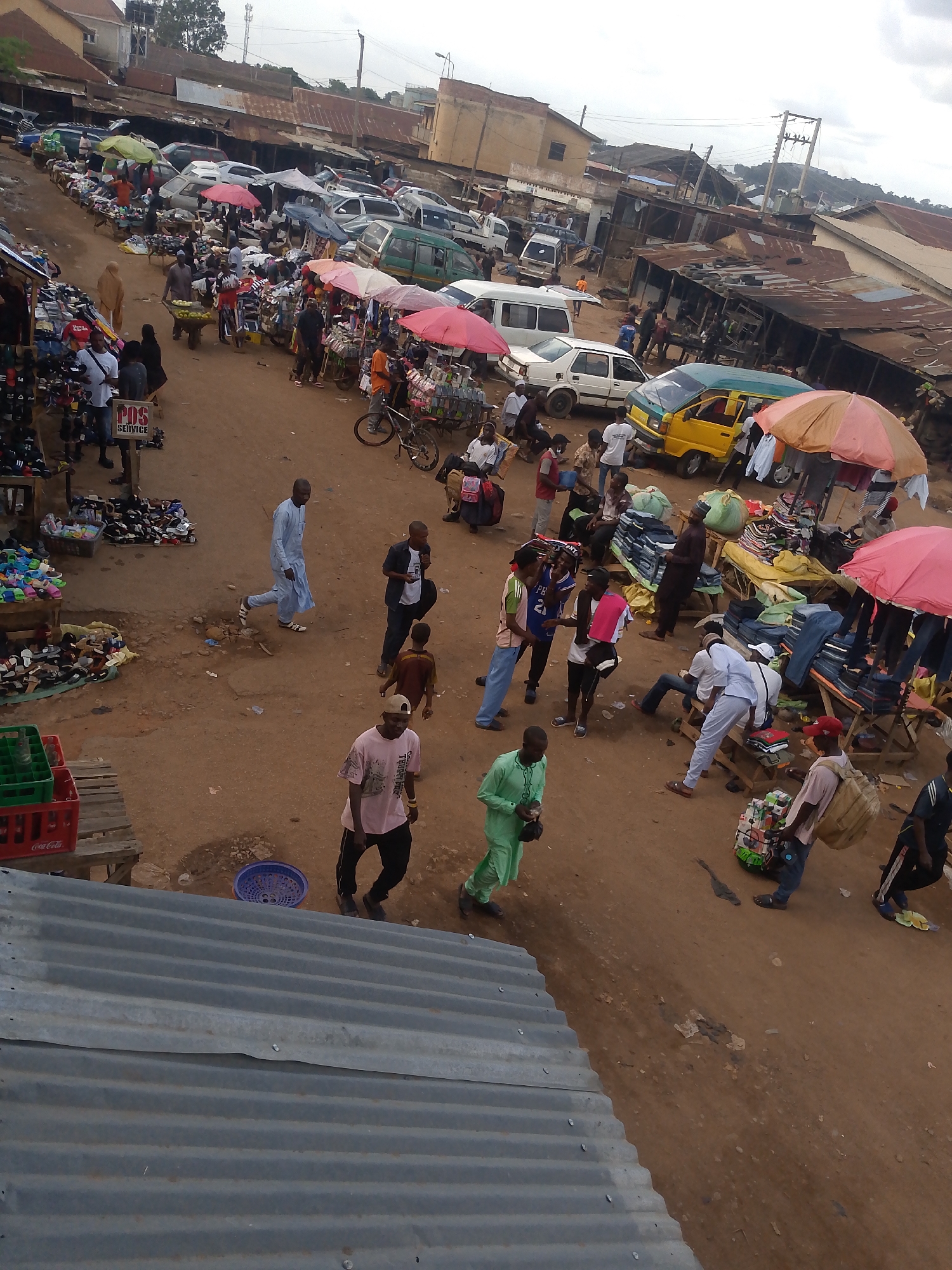 This photograph depicts a bustling street market in Bukuru, Nigeria. The scene is viewed from an elevated perspective, looking down upon a crowded area filled with people, vehicles, and numerous stalls. The ground is primarily a reddish-brown dirt.
People of varying ages and attire are present, some walking, others gathered around vendors. There are individuals wearing traditional clothing, while others are in more casual attire. A variety of goods are displayed for sale beneath colorful umbrellas or on tables and carts. These items appear to include clothing, shoes, and assorted merchandise.
Vehicles, including cars and vans, are parked haphazardly along the street, adding to the congestion. Buildings with corrugated metal roofs line the periphery of the market area. Overhead, a cloudy sky provides diffuse lighting. Visible in the distance are electricity poles and other urban structures. The overall impression is of a lively and active marketplace in a densely populated urban setting.