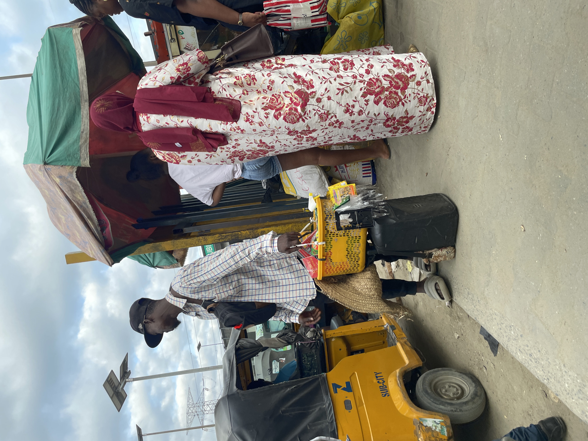 The image captures a street scene in Lagos, Nigeria, likely near a transportation hub or market. In the foreground, a man wearing a plaid shirt, a black baseball cap, and dark pants stands beside a yellow tricycle, also known as an "okada" or "keke." He is carrying a woven straw bag on his left shoulder and holding a yellow plastic basket filled with various items, including what appear to be packaged goods and some loose food items. Beside him, a black rolling suitcase stands on the ground.
To the right of the man, a woman is seen from behind, wearing a long, white dress with a prominent floral print of red roses and a matching red hijab. Another woman, wearing a white t-shirt and denim shorts, is visible in the middle ground, to the left of the woman in the floral dress. They are standing under a makeshift canopy supported by yellow poles, which appears to be a stall or shelter. The canopy has a faded green and red fabric.
In the background, a transmission tower and solar-powered streetlights are visible against a cloudy sky, indicating an urban setting. The ground is a mix of paved concrete and dirt. The yellow tricycle has "SUB-CITY" written on its side. The overall atmosphere suggests a busy day with people engaged in commerce or transport.