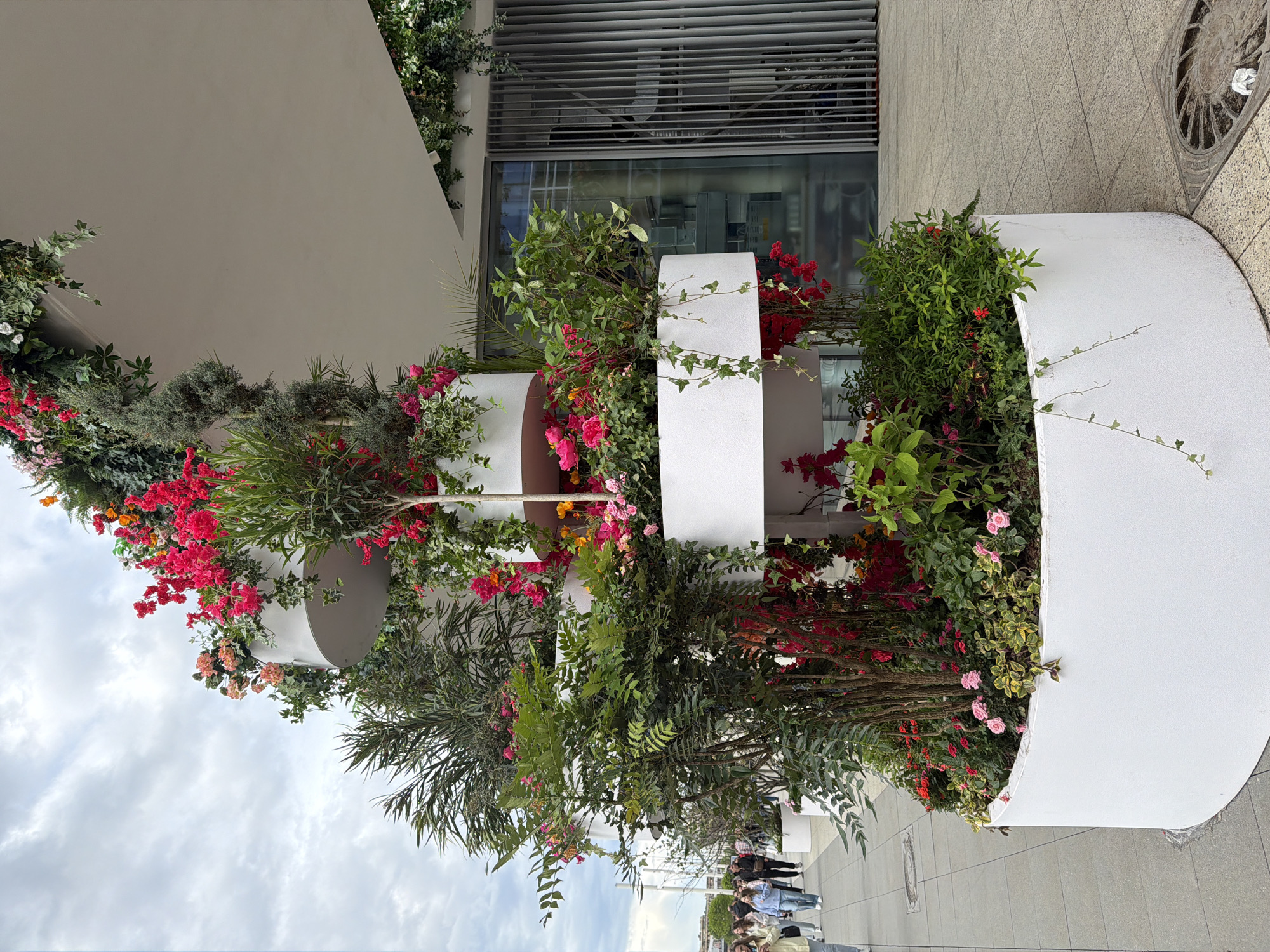 The image shows a tiered, white planter filled with a variety of flowering plants and greenery, possibly located in Moscow, Russia, based on the architectural style and the presence of Russian-speaking individuals in the distance. The planter has a curved, cylindrical design with multiple levels, creating a cascading effect of vegetation. Red, pink, and orange flowers are prominent, mixed with various types of foliage.
The planter is situated on a paved walkway next to a building with a modern facade, including large windows and a metallic-looking structure above. In the background, a group of people can be seen walking along the sidewalk. The sky is overcast with a mix of clouds.