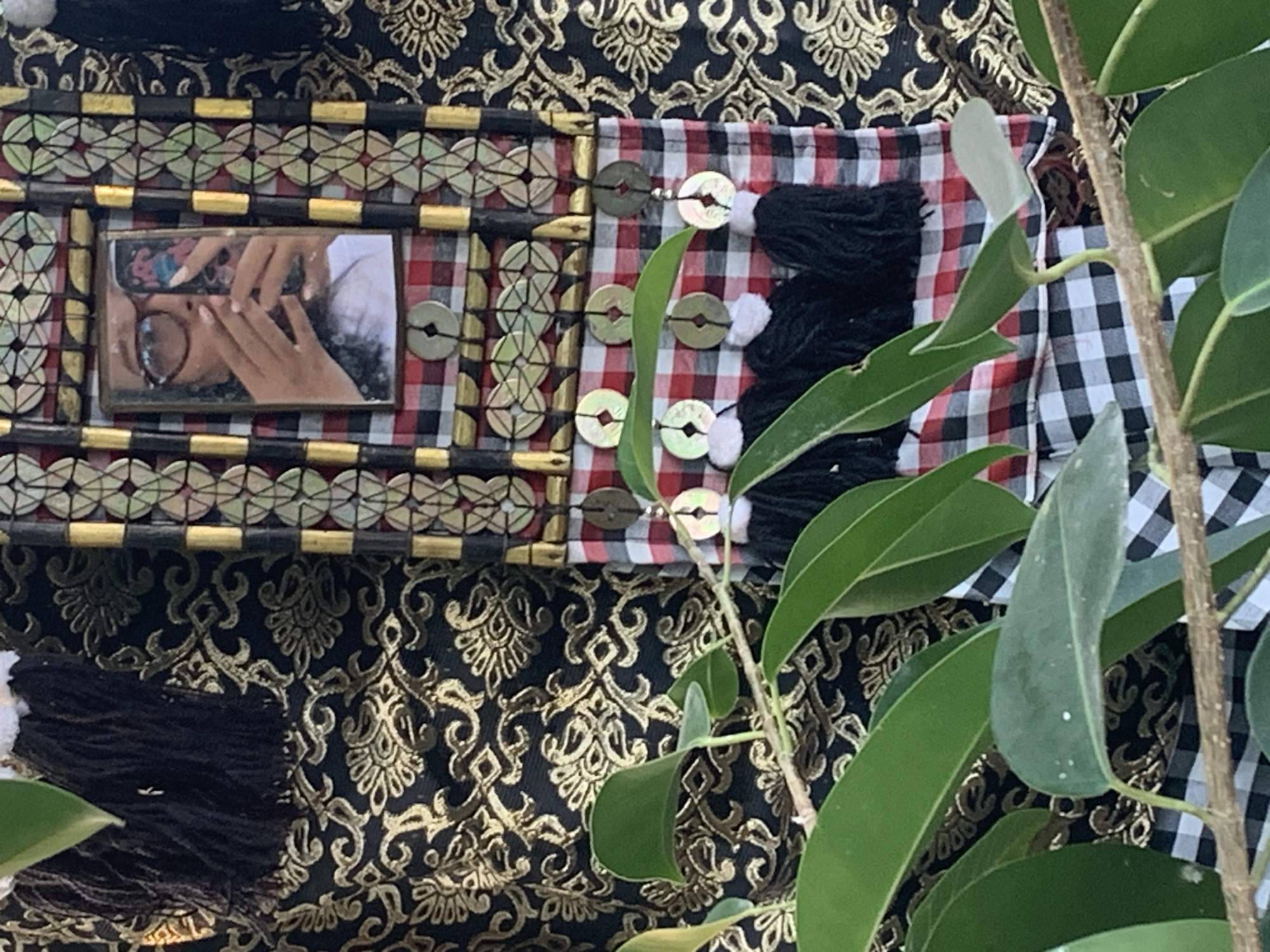In Sukawati, Indonesia, a detailed close-up shot reveals a decorative fabric adorned with intricate golden patterns on a black background. A central vertical element features a framed reflection of a person holding a phone, wearing glasses, and taking a selfie. This frame is bordered by black and gold embellishments and a checkered red and white fabric. Below the frame, strings of coins and black tassels are suspended from the checkered fabric. In the foreground, lush green leaves and branches partially obscure the view of the decorative fabric.