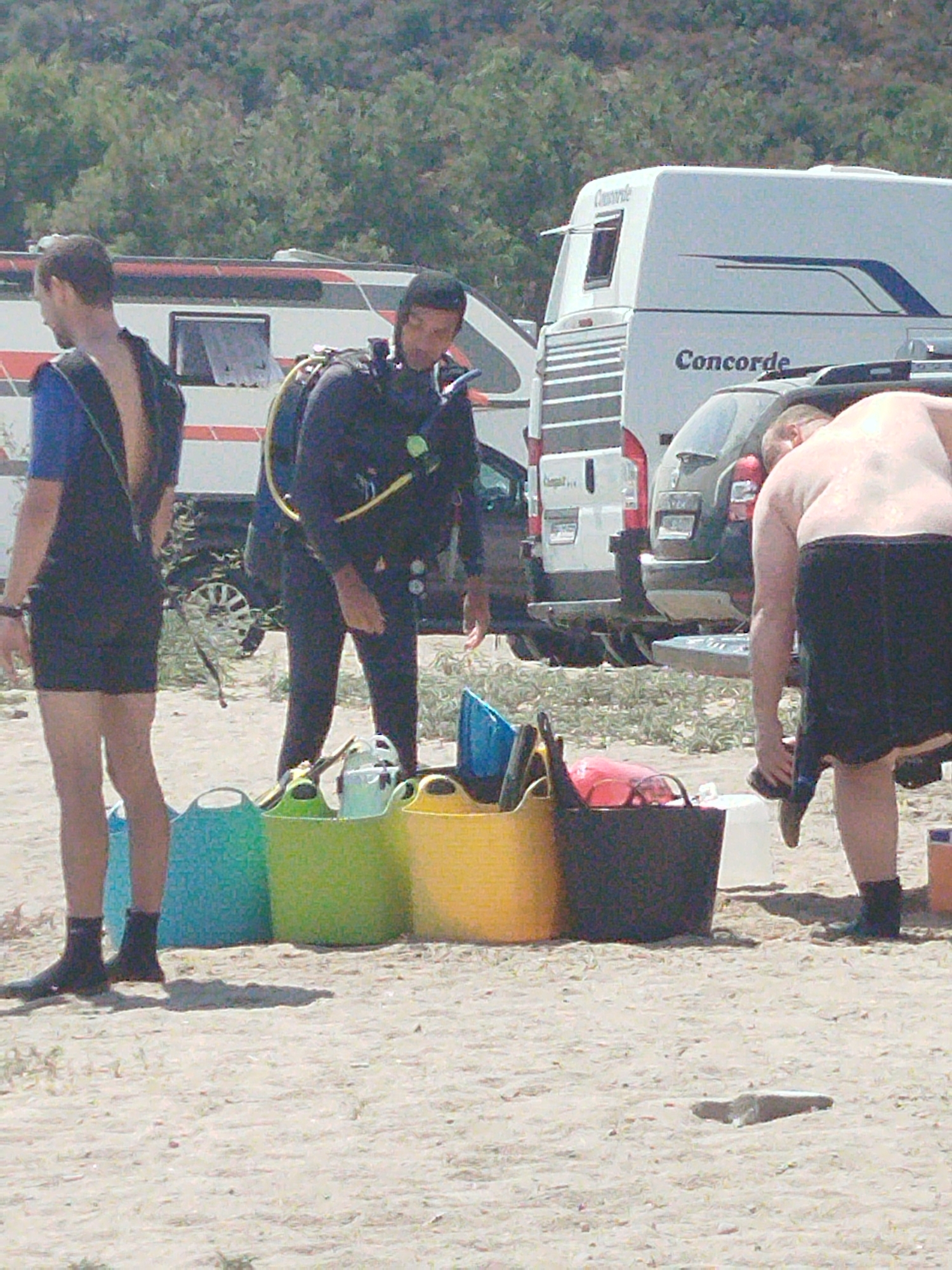 Three individuals are present on a sandy beach in Sithonia Municipal Unit, Greece, with a white recreational vehicle and a dark colored vehicle in the background. The individual on the left, a male, is wearing a dark blue wetsuit. The individual in the center, also male, is wearing a full black wetsuit with scuba gear attached, including a yellow hose and a black tank. The individual on the right, a male with a visible tan line, is wearing black shorts and is in the process of putting on a black shoe. In the foreground, several large plastic buckets are arranged, in shades of blue, green, and yellow, with some equipment within them. The environment is characterized by sand, sparse vegetation, and a bright, sunny atmosphere.