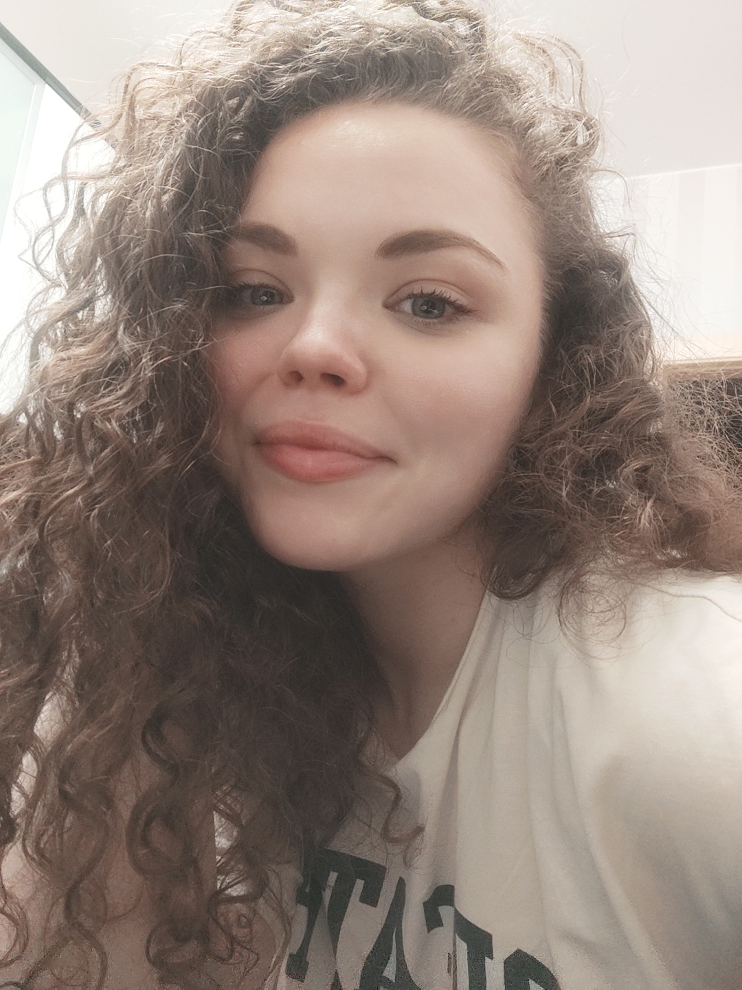 A young woman with curly brown hair is looking at the camera. She is wearing a white t-shirt with dark green letters. The background is out of focus, but appears to be an indoor setting.