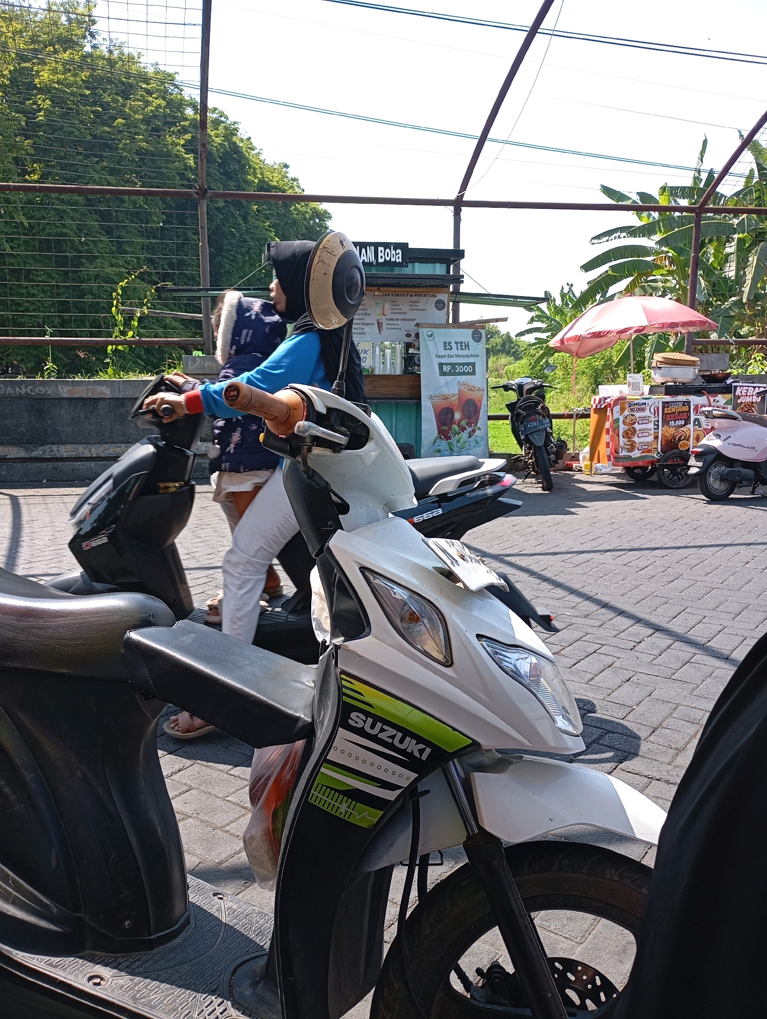 In Kalisampurno, Indonesia, a woman wearing a blue shirt, white pants, and a black hijab rides a black scooter. She is accompanied by a child in a blue jacket with white trim. The scooter has a black seat and appears to be parked on a brick-paved area. In the background, there are food stalls selling items like dim sum, kentang, and kebab. A pink scooter is also visible. A large tree with green foliage is to the left of the frame. The sky is bright and appears to be a clear day.