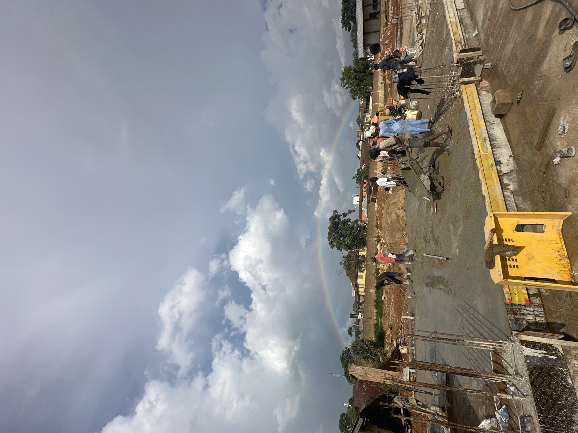 The image depicts an outdoor construction site in Jos, Nigeria, under a dramatic sky with a visible rainbow. Several men are engaged in construction work on a concrete foundation. In the foreground, rebar is visible, suggesting the early stages of construction. A yellow construction barrier is placed on the left side of the frame. A group of men are working with a cement mixer and wheelbarrows on the wet concrete surface. Further in the background, more structures and trees are present. The sky is filled with dark, heavy clouds, with patches of lighter sky, and a distinct rainbow arcs across the scene. The ground is wet, indicating recent rain, which likely caused the rainbow.