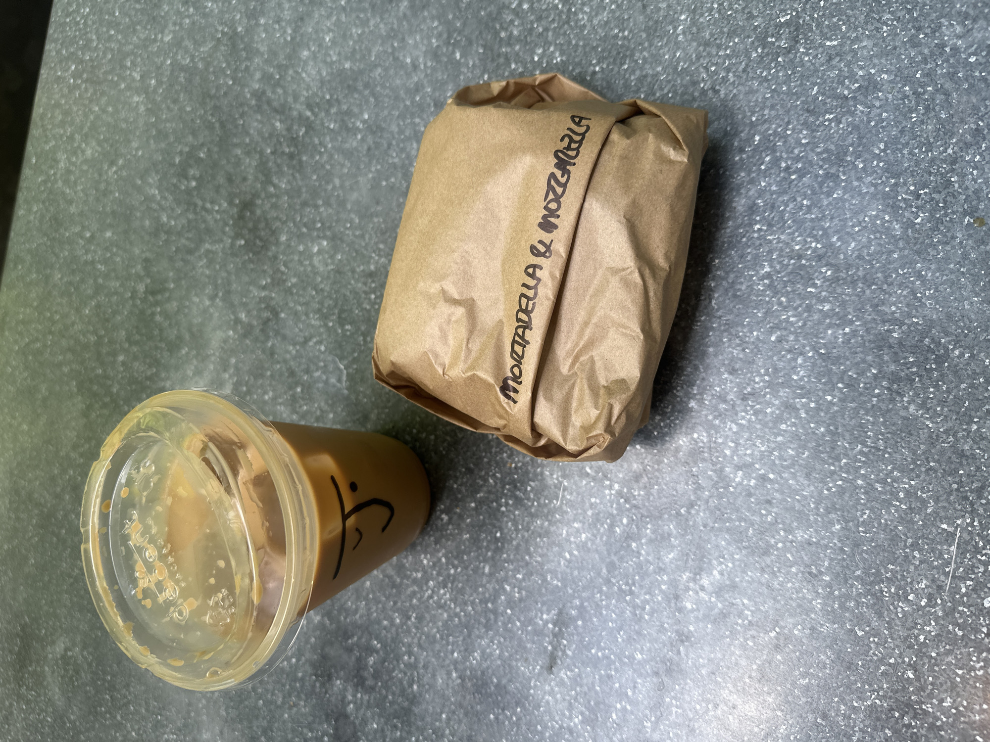 A partially consumed iced coffee in a clear plastic cup, labeled with a graphic design, and a Mortadella & Mozzarella sandwich wrapped in brown paper are positioned on a speckled gray surface. The brown paper of the sandwich wrapping is wrinkled. The scene seems to be indoors.