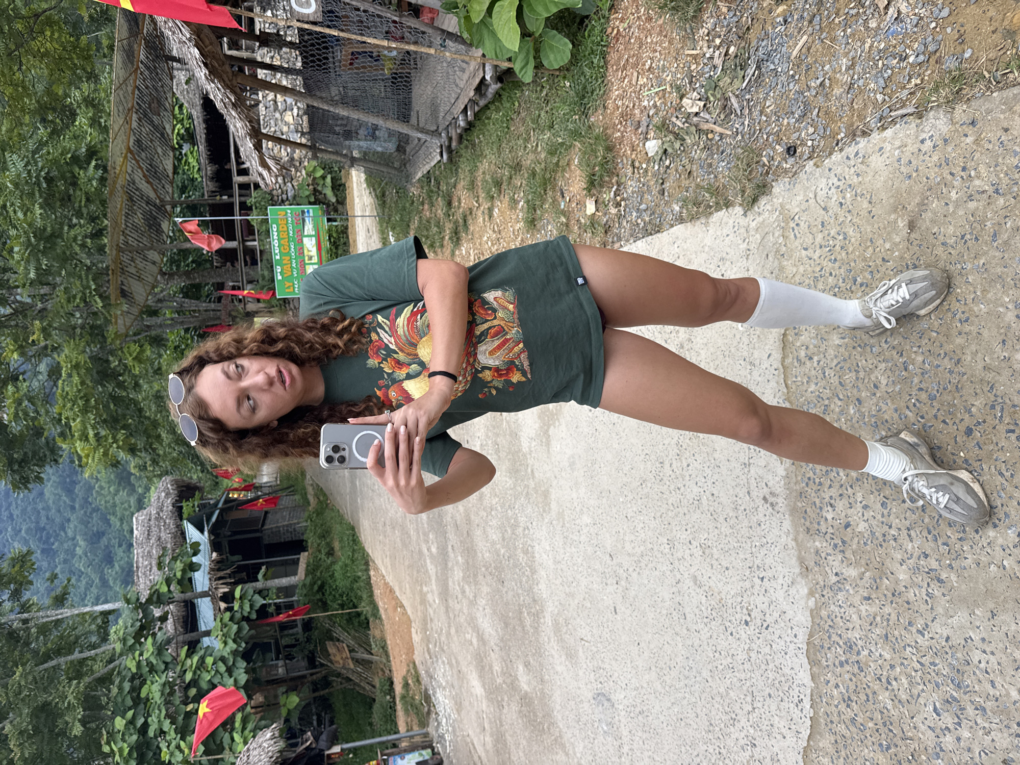 A young woman with curly brown hair, wearing a green t-shirt with a bird graphic, white knee-high socks, and grey sneakers, is taking a selfie. She is standing on a paved path in a rural area. The path is bordered by greenery and some rustic buildings with thatched roofs. Red flags are visible in the background, suggesting a location in Vietnam. A sign with Vietnamese text is also present. The woman is looking at her phone, which she holds in her right hand, and has a relaxed expression. The scene appears to be outdoors, during daylight. The background shows a lush, hilly landscape.