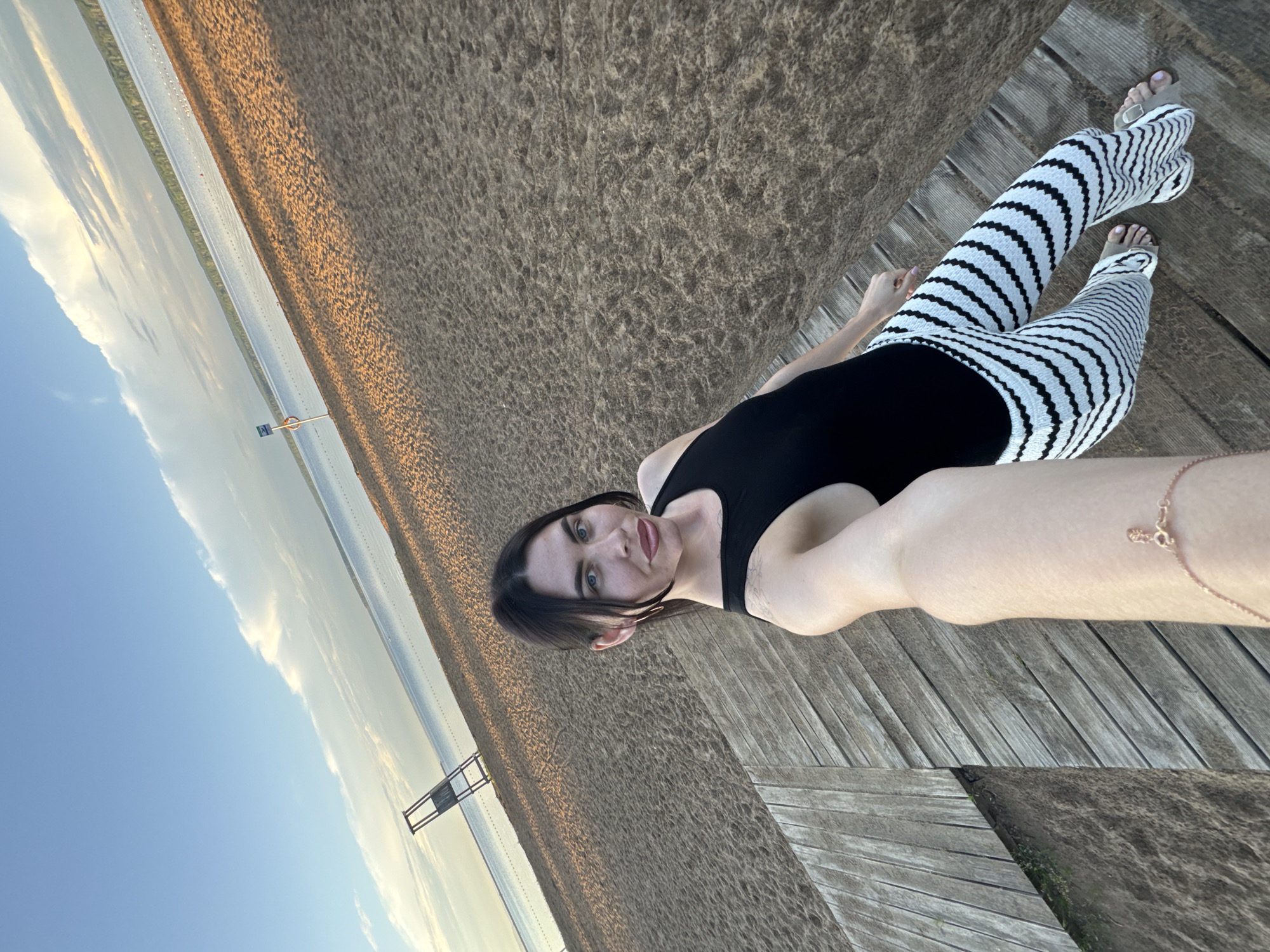 A woman stands on a wooden walkway at a beach, holding a camera to take a selfie. She is wearing a black tank top and black and white striped pants. The beach is sandy, with a body of water in the background. The sky is blue with some clouds. The location is the Kama River beach in Chaikovsky, Russia.