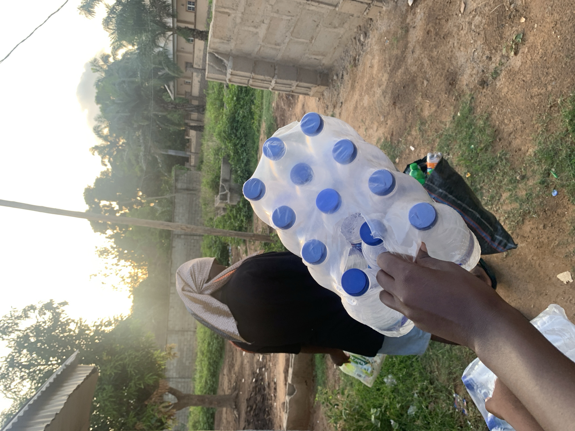 The image shows a person removing a water bottle from a package of bottles. The person is wearing a dark shirt and a head covering. Another person stands nearby. The setting appears to be outside near a dirt road. The environment includes trees, a building made of concrete blocks, and other vegetation. Based on the location provided, this scene is most likely in Keffi, Nigeria.