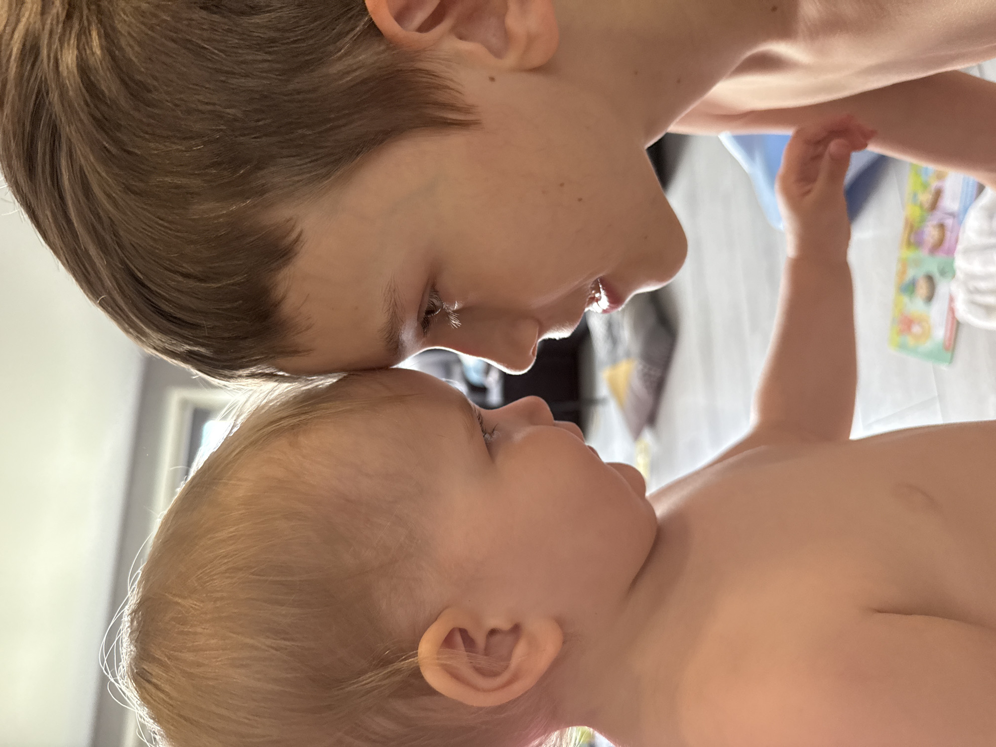 The image shows a close-up of two young children, likely siblings, facing each other. The older child, with brown hair, is looking down with their head tilted. Their mouth is slightly open, revealing teeth. The younger child, with blonde hair, is looking up, their eyes closed. Their skin is soft and smooth, and their ear is visible. The children are positioned forehead-to-forehead, suggesting an intimate moment of connection. The lighting is bright, hinting at daytime. In the background, blurry objects include what appears to be a book or toy with colorful illustrations and possibly some fabric. The scene appears to be indoors, likely in a home, with a hint of a light-colored floor and a doorway or window in the distance. The overall emotion conveyed is one of tenderness and affection between the siblings. No visible text is present in the image.