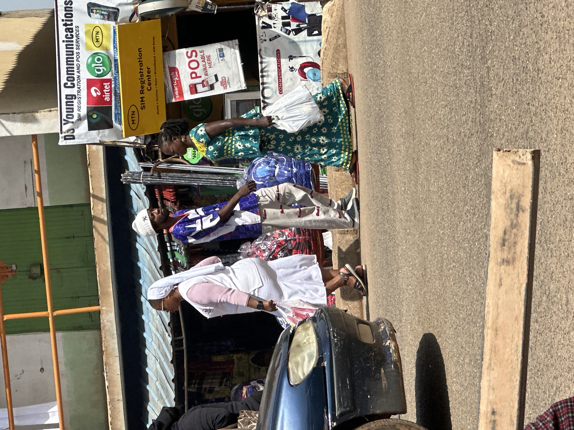 The image captures a street scene in Bukuru, Nigeria, under a bright, sunny sky. A dark blue car is partially visible on the left side of the frame, parked or moving on a paved road. In the center, three individuals are walking. A woman in a pink top and a white head covering, wearing sandals, carries a white plastic bag. Beside her, a man in a white beanie and a blue and white patterned shirt with grey pants, also in sandals, walks in the same direction. To the right of the man, a woman in a vibrant green and yellow patterned dress, also wearing sandals, carries a white plastic bag. They appear to be moving away from the camera.
Behind the pedestrians, a row of shops line the street. One shop has a prominent yellow sign with "MTN SIM Registration Center" written in black text. Above this, a larger white banner displays "DS Young Communications SIM REGISTRATION AND POS SERVICES" along with logos for Airtel, Glo, and MTN, as well as images of cell phones. Another sign below the MTN banner advertises "smartcash POS AVAILABLE HERE." Goods are displayed behind the shops, including clothing on racks and various packaged items. The environment suggests a busy commercial area.