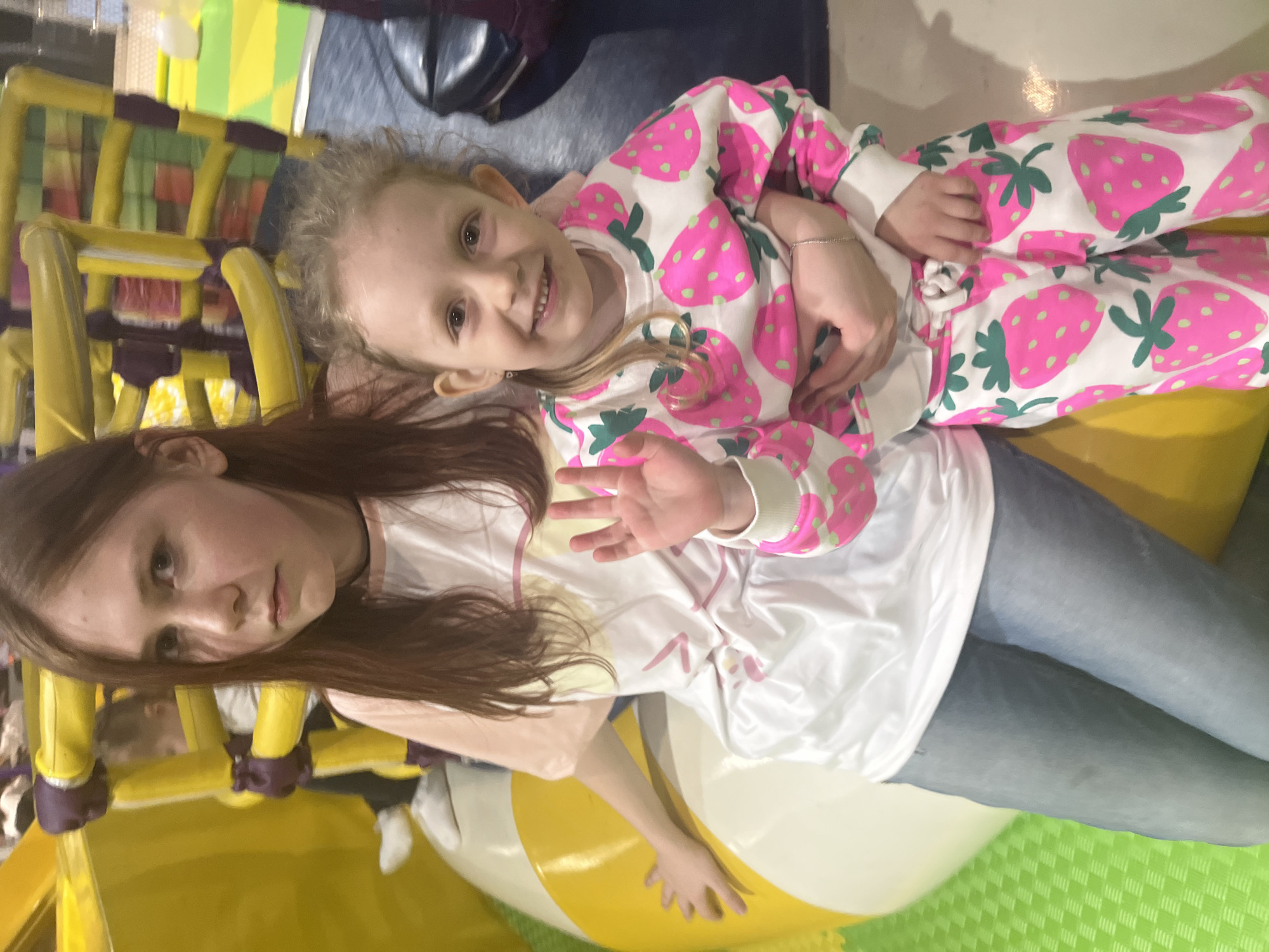 Two Caucasian females are indoors at a recreational facility, likely a children's play area. The older female has long brown hair, is wearing a white t-shirt, light pink top, and blue jeans. The younger female has blonde hair and is wearing a white pajama set with pink strawberries on it. The younger female is resting on the older female's chest. They are both smiling. The background includes yellow and purple play structures. The location cannot be determined.