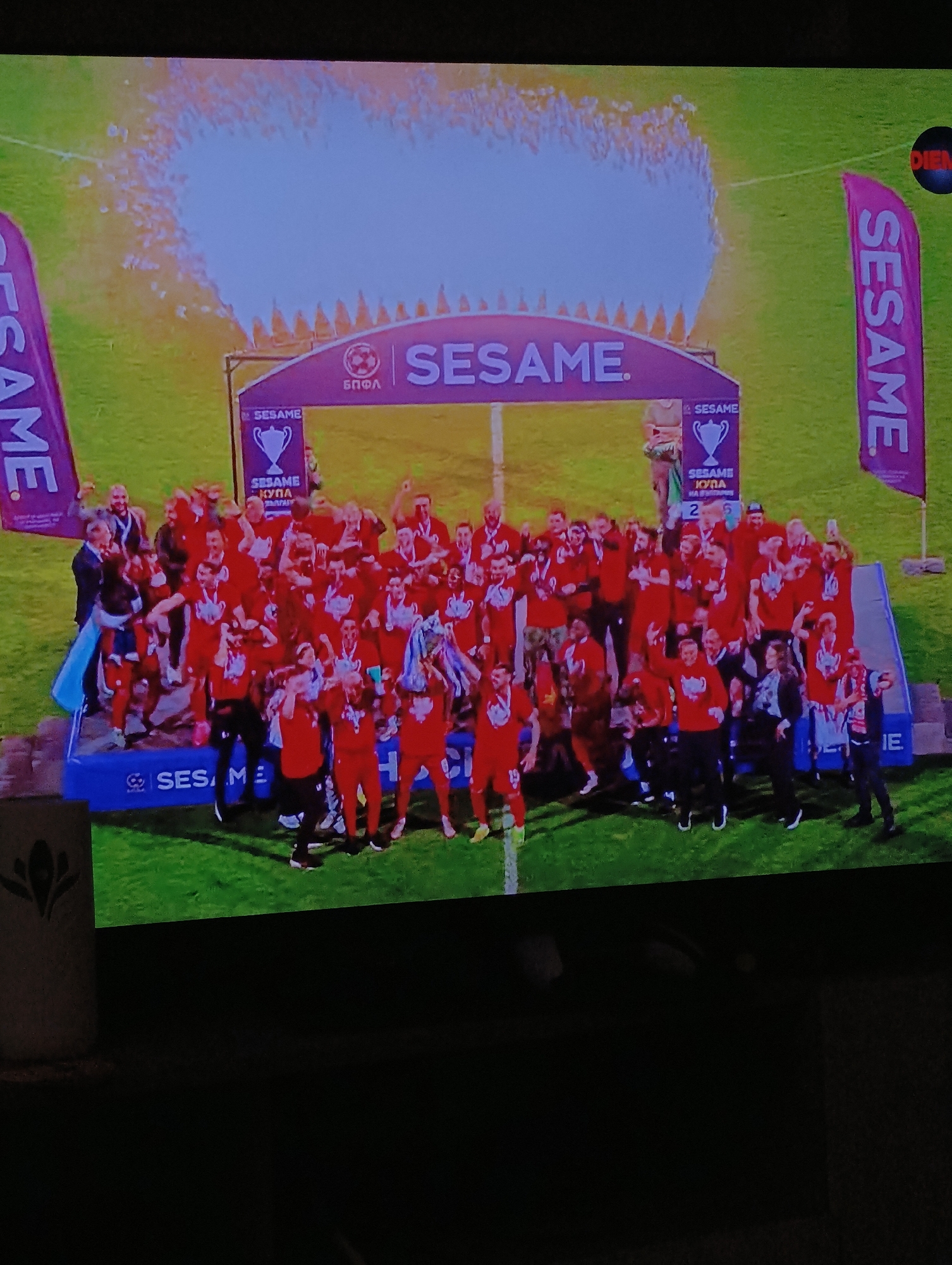 The image captures a celebratory moment on what appears to be a soccer field, under bright lighting or sunlight, with traces of celebratory smoke above an archway. A large group of people, presumably a winning team, is clustered on a small platform or stage, many wearing red jerseys. They seem to be jubilant, holding their arms up in triumph and interacting with each other in a celebratory manner. Behind them, there is a large archway with the word "SESAME" printed on it. Tall purple flags with the same word "SESAME" are positioned at either side of the stage. The environment is a green field. In the bottom left corner, there is a cropped portion of what looks like a patterned vase or container. The overall scene suggests a victory celebration at a sporting event. No specific location details such as the city of Plovdiv, Bulgaria can be determined from the image.