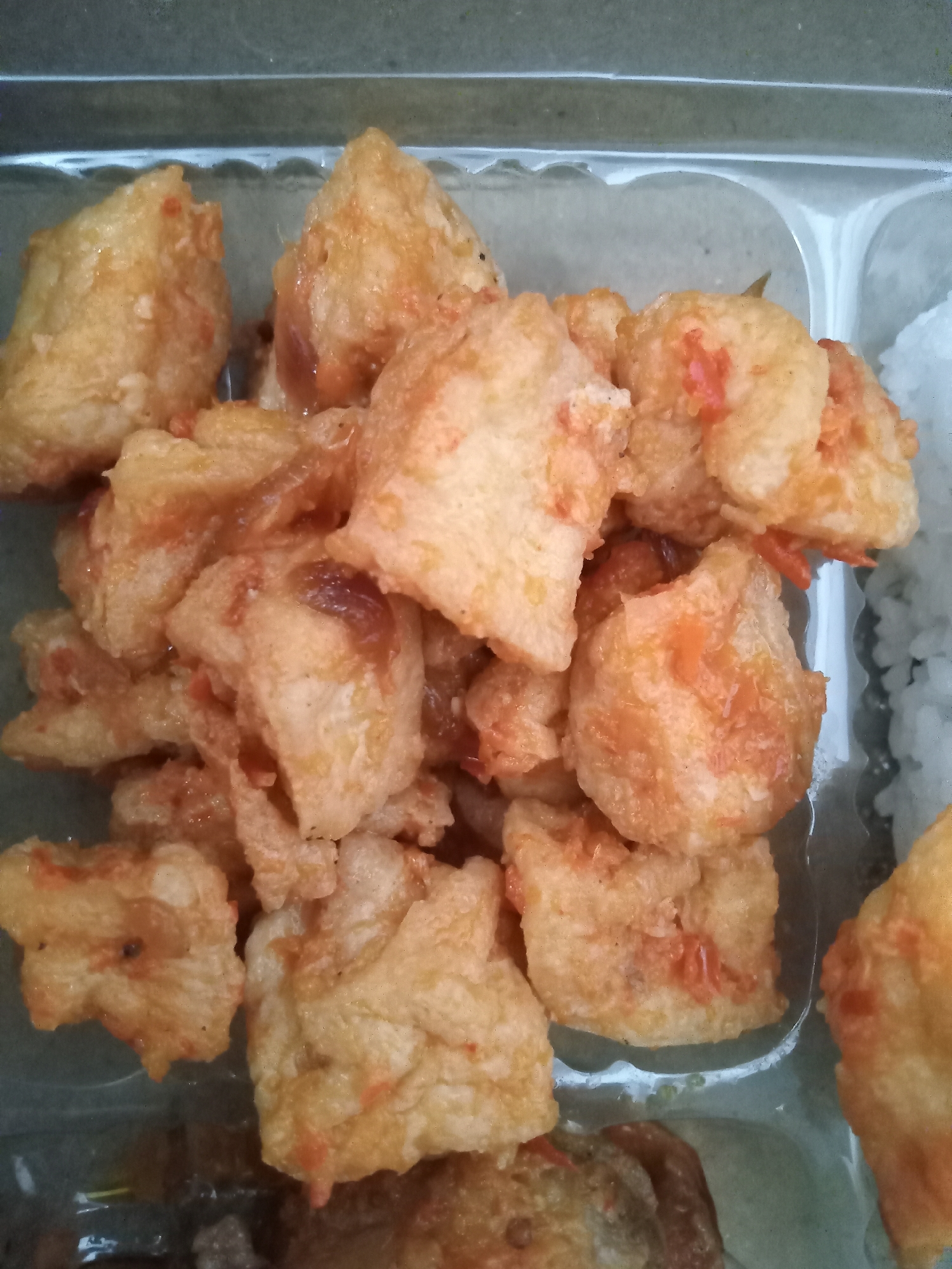 This image shows a close-up view of fried tofu in a plastic container, likely from a food stall in Simo, Indonesia. The tofu pieces are bite-sized, golden brown, and coated in a reddish sauce with visible bits of chili or tomato. They are piled together, obscuring the container's bottom. To the right of the tofu, a small portion of white rice is visible. The background is out of focus, showing the interior of what appears to be a generic food stall. The lighting is direct, causing some reflections on the plastic container.