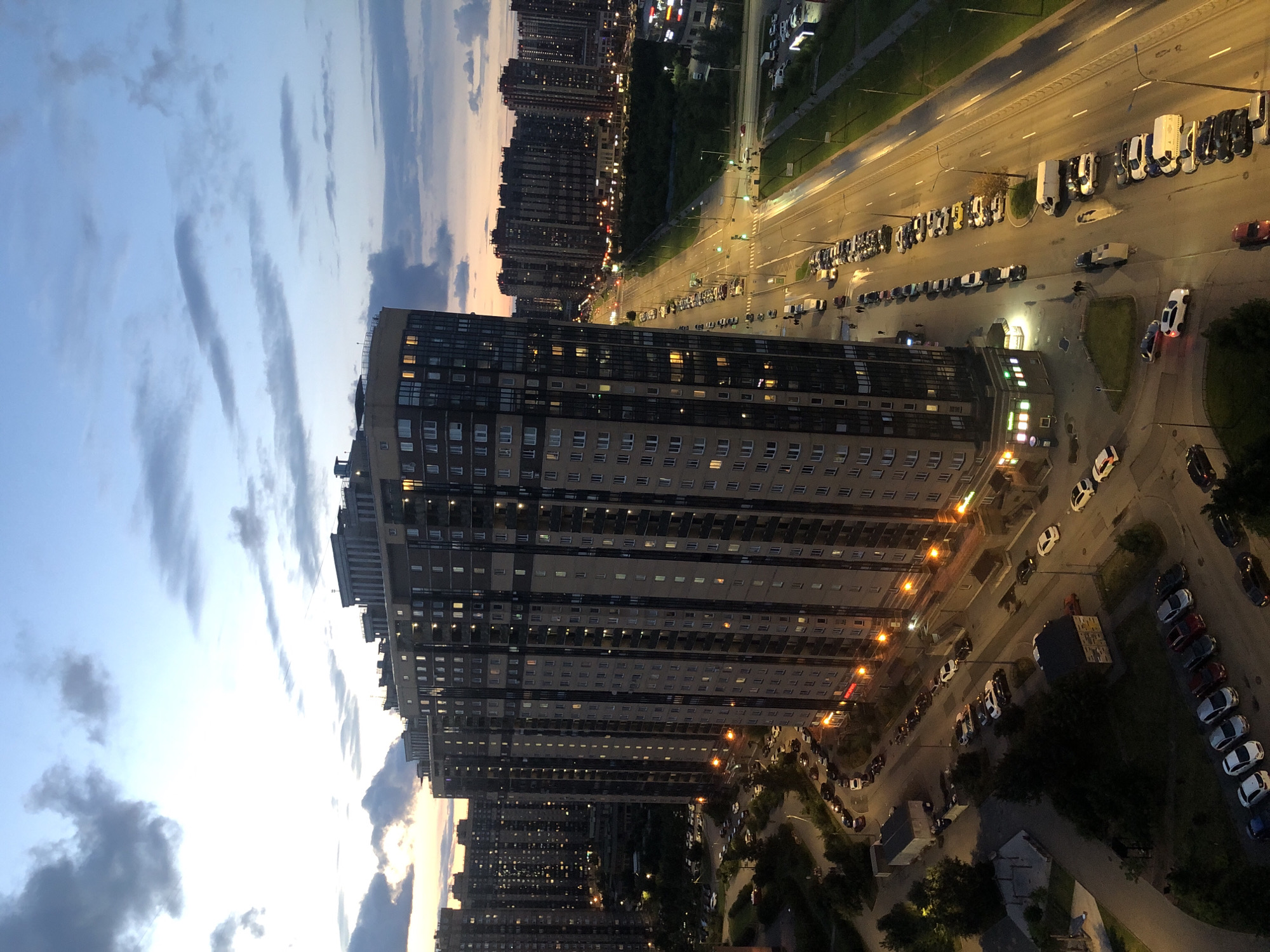 An aerial, eye-level view captures a dense urban environment at dusk. The foreground is dominated by a towering, multi-story residential building with numerous illuminated windows, indicating it is occupied. Adjacent to this building is a street lined with parked cars and a few pedestrians. The street stretches into the distance, flanked by more residential buildings and illuminated by streetlights. The sky above is a gradient of blue and orange, with scattered clouds, suggesting the sun has recently set. The overall scene depicts a typical residential area within a city during the evening hours.