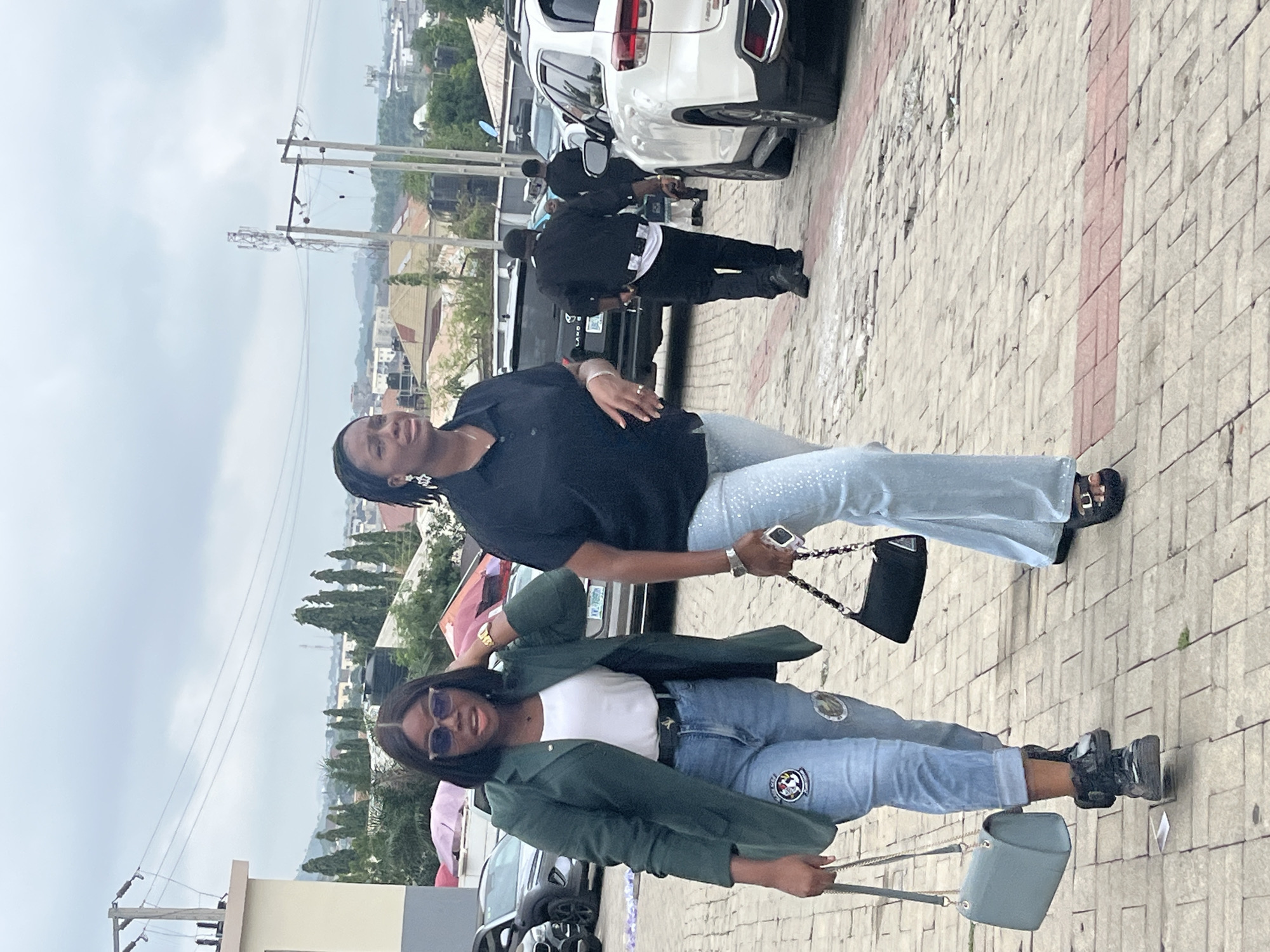 Two women walk side-by-side on a paved sidewalk in Abuja, Nigeria. The woman on the left wears a dark green blazer, a white top, and light-wash jeans adorned with patches. She carries a light blue handbag in her left hand and has her right hand near her head, adjusting her sunglasses. The woman on the right wears a black collared shirt and light blue, shimmering bell-bottom pants. She carries a black handbag with a chain strap in her right hand and appears to be looking forward. Behind them, several vehicles, including a white SUV and a black SUV, are parked. Power lines and some buildings are visible in the background, with a hazy, overcast sky overhead.