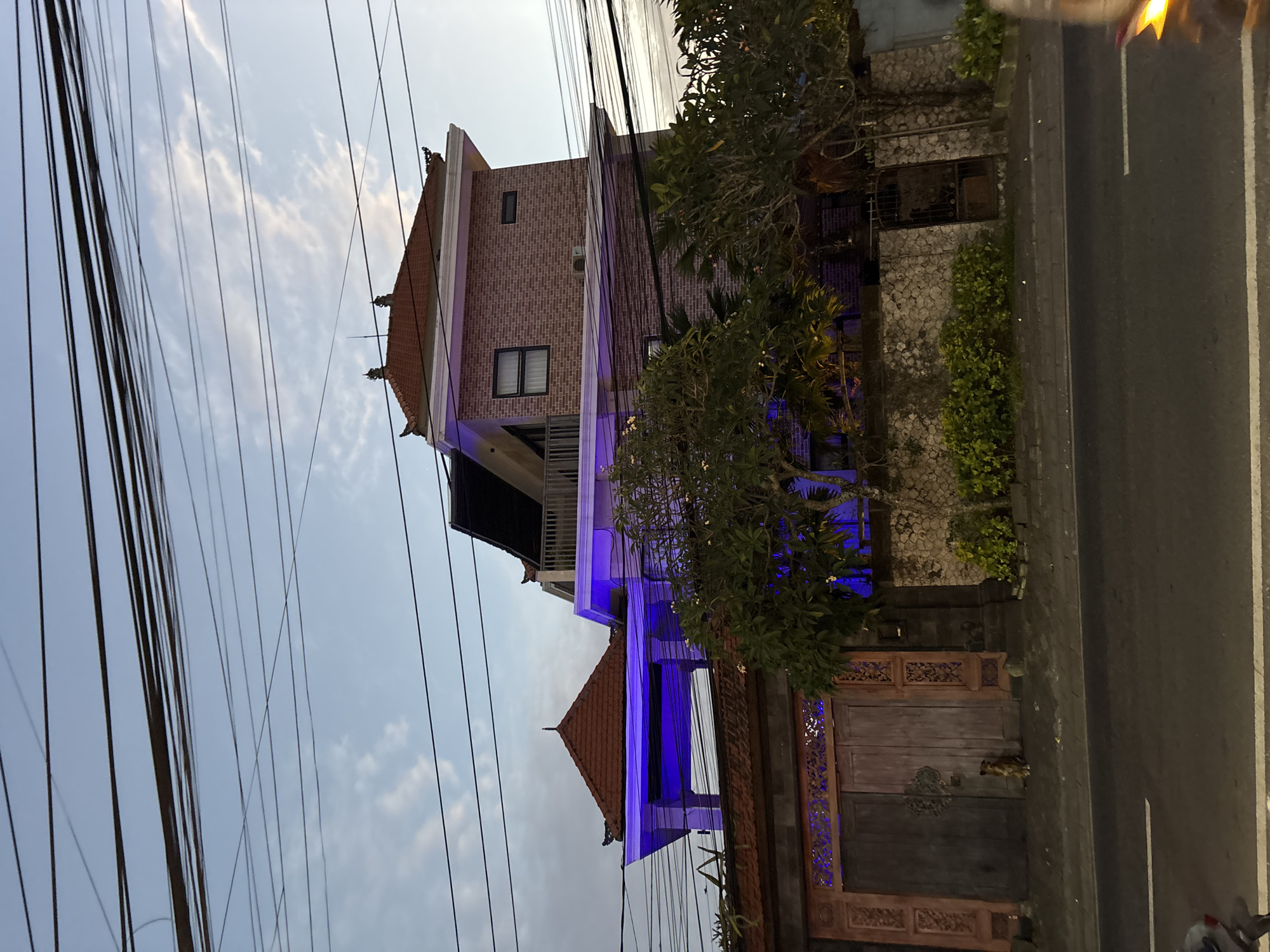 A nighttime view shows a three-story building illuminated with blue lights. The building's exterior is constructed of reddish-brown brick, with a roof of similar color and style. A small balcony is visible on the second floor.
The building is situated next to a road, and several power lines cross in front of the scene. Vegetation, including trees and shrubs, partially obscures the building. There are wooden gates or doors near the base of the building, decorated with ornamental carvings.
The image was taken in Kuta Selatan, Indonesia. No people are visible in the frame.