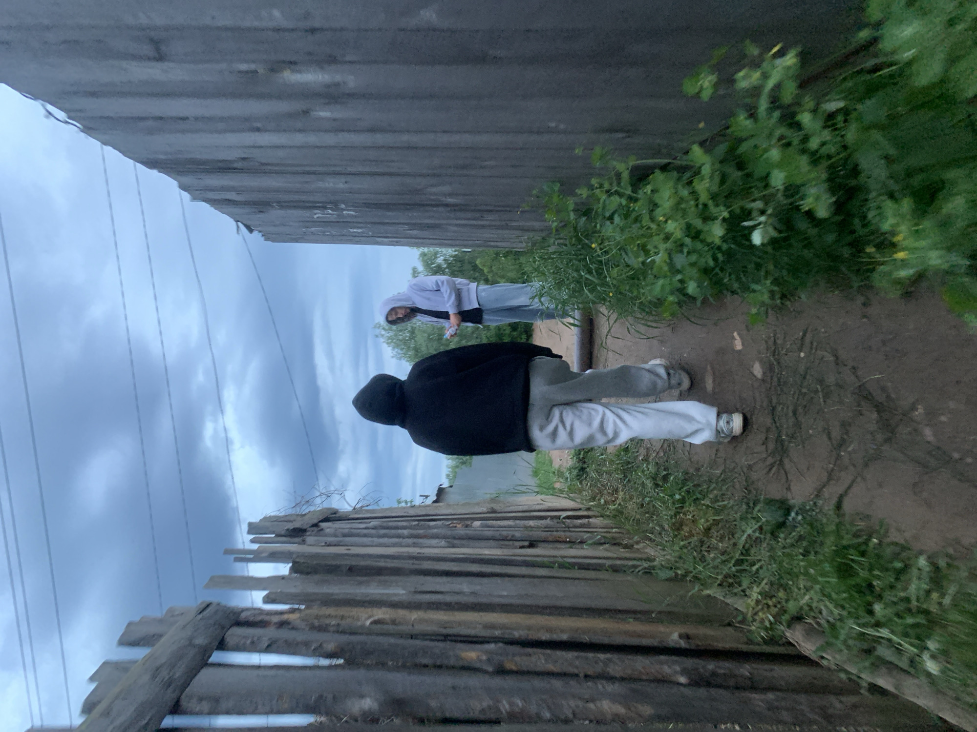 The image shows a path between two wooden fences under an overcast sky. A person wearing a black hoodie and gray sweatpants walks away from the camera. Another person in a light purple hoodie and light-colored pants stands further down the path. Green vegetation grows along the sides of the path and fences. The image was taken in Chaikovsky, Russia.
