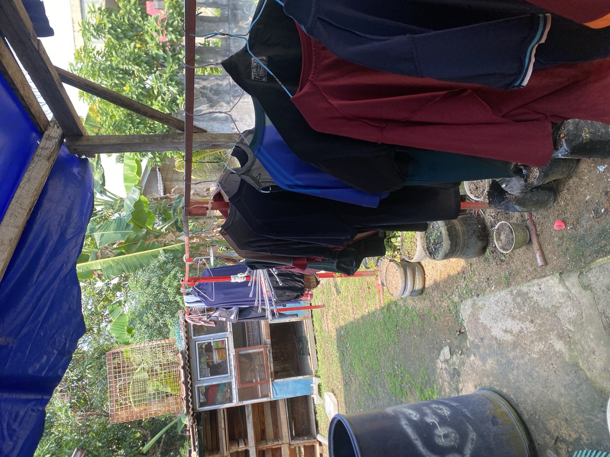 This is an outdoor scene, likely a backyard in a residential area of Batam, Indonesia. In the foreground, a large dark blue cylindrical object, possibly a water tank or bin, stands on a concrete surface. The surface is damp and shows signs of wear. To the right, several potted plants in various containers, some made of recycled materials, are arranged on the ground, which is a mix of dirt and gravel.
Further back, a clothesline made of red metal poles holds numerous items of clothing hanging on hangers. The clothes include t-shirts and other garments in various colors such as black, dark blue, maroon, and grey. To the left of the clothesline is a structure that appears to be a coop or shelter, constructed from wood and metal mesh, with several compartments. One compartment has a window with a label that reads "ARIS". A rusty orange metal cage is placed on top of this structure.
The background reveals lush green vegetation, including banana trees and other leafy plants, indicative of a tropical climate. In the far background, a concrete wall and buildings can be seen, suggesting the presence of neighboring houses. A blue tarp is stretched over a wooden frame above the clothesline, providing some shade. The overall impression is of a functional, somewhat rustic outdoor space. No people are visible in the image.