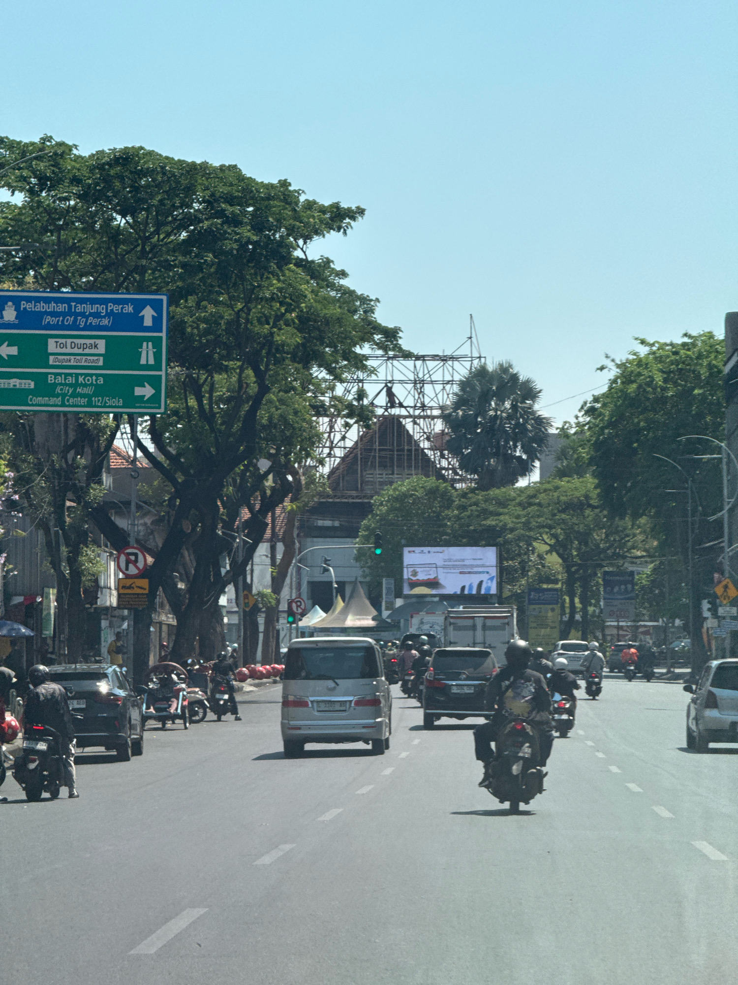 This is a street-level, eye-level, medium shot of a busy road in Surabaya, Indonesia, likely taken from a vehicle. The foreground is dominated by the asphalt road with white lane markings, and several vehicles are visible, including cars and motorcycles, with riders mostly wearing helmets. A silver van is in the center lane, and behind it, a black SUV is also visible. Several motorcycles are navigating the traffic.
On the left side of the road, a blue directional signpost is prominent. It indicates "Pelabuhan Tanjung Perak (Port Of Tg Perak)" with an upward arrow, "Tol Dupak (Dupak Toll Road)" with a leftward arrow, and "Balai Kota (City Hall) Command Center 112/Siola" with a rightward arrow. Behind the sign, mature trees with dense green foliage line the street. A red and white circular "no parking" sign is also visible on the left.
Further down the street, in the background, a large digital billboard displays an advertisement. More trees and buildings are visible, with scaffolding on one of the structures, suggesting ongoing construction. The sky is clear and blue, indicating a sunny day. The overall scene depicts a typical urban environment in Surabaya, with active traffic and local signage.