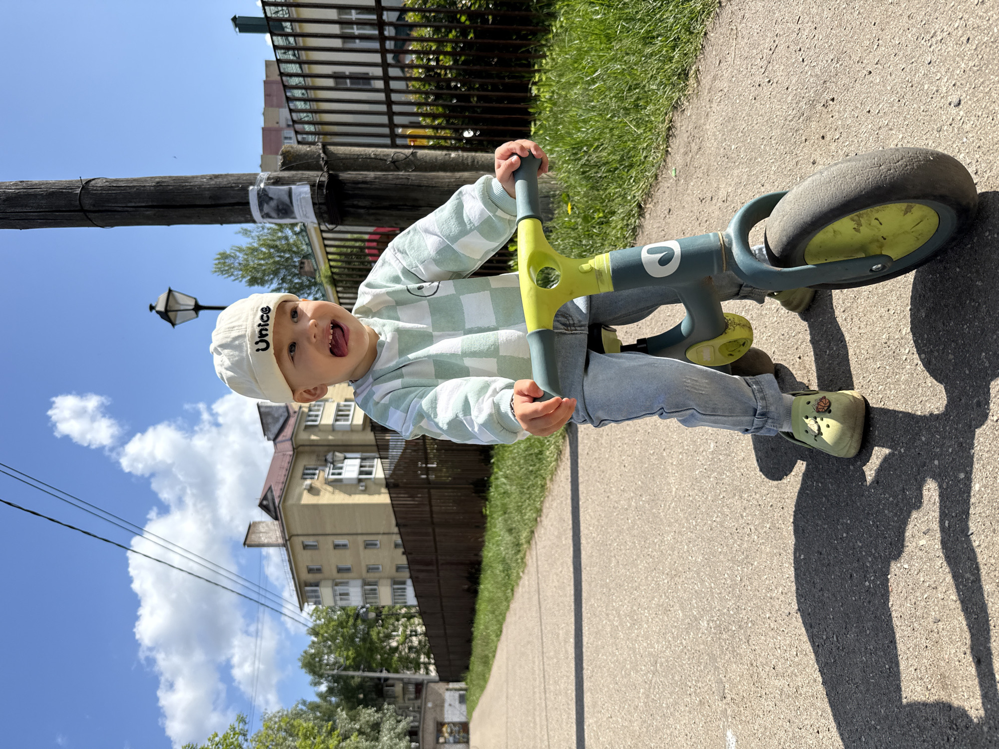 A young child, appearing to be a toddler, is depicted outdoors on a sunny day in Rusanovo, Russia. The child is wearing a white baseball cap with "Unice" written on it, a light green and white checkered long-sleeved hoodie, light blue jeans, and olive green Crocs. They are seated on or pushing a grey and lime green balance bike, holding onto its handlebars. The child's face is expressive, with an open mouth and tongue slightly extended, conveying a playful and joyful emotion.
The scene is set on a paved path or sidewalk, with a strip of green grass and a dark fence running alongside it. In the background, several multi-story buildings with residential-style architecture are visible under a bright blue sky with scattered white clouds. A tall wooden utility pole with a street lamp and overhead power lines is prominent to the left. A small, unreadable paper is taped to the wooden pole. Strong shadows on the ground indicate bright daylight, suggesting either morning or afternoon.
