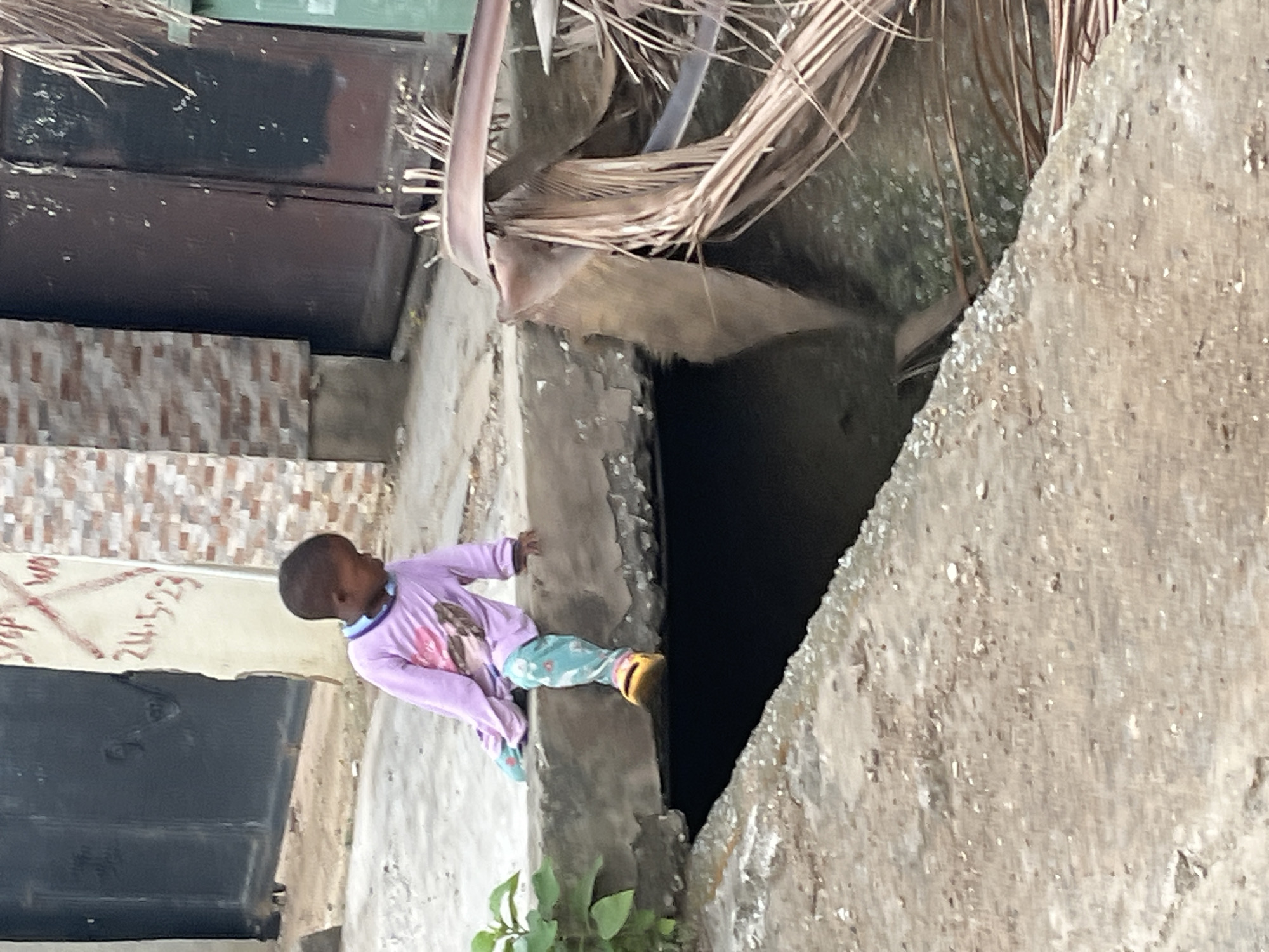 A young child wearing a lavender long-sleeved shirt and patterned blue pants is sitting on the edge of a concrete structure. The child appears to be looking downwards into a dark opening. The surrounding environment suggests an urban setting with a combination of concrete and decaying organic material. Based on the prompt, the location is Abuja, Nigeria.