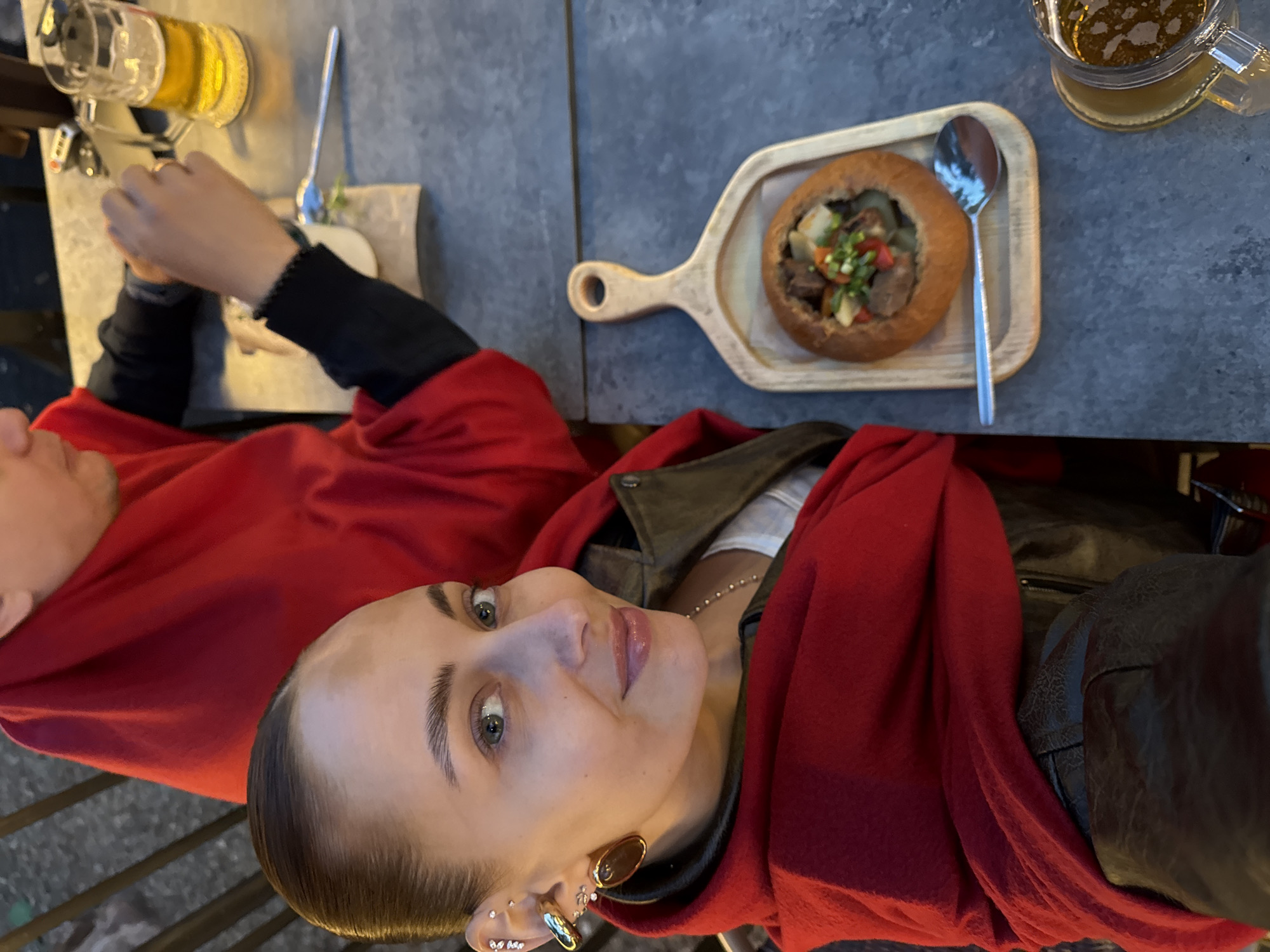 This image captures a moment of dining in Izhevsk, Russia, likely during the evening due to the artificial lighting illuminating the subjects.
The main subject is a woman in the foreground, seen from a high angle looking upwards towards the camera. She has slicked-back dark hair, light makeup, and multiple earrings on her left ear, including a large gold oval one and several smaller studs. She is wearing a red top or scarf over a dark leather jacket and a white or light-colored top underneath, visible at the neckline. Her expression is neutral to slightly pleasant.
Beside her, another person is partially visible, wearing a red top and a black long-sleeved shirt. This person's hands are visible near a glass of beer, appearing to be engaged with something off-camera or just below the frame.
On the dark, textured tabletop between the two individuals, there are two drinks and a meal. One glass of amber-colored beer is near the partially visible person. The main meal consists of a stew served inside a hollowed-out bread bowl, placed on a light wooden paddle-shaped board. The stew contains pieces of meat, potatoes, and other vegetables, garnished with fresh green herbs. A silver spoon rests beside the bread bowl. Another glass of beer is visible in the background on the table.
The setting appears to be an outdoor or semi-outdoor cafe or restaurant, indicated by the table and the metallic railing visible in the foreground. No specific location cues beyond the general dining environment are present in the image itself, and no text is visible.