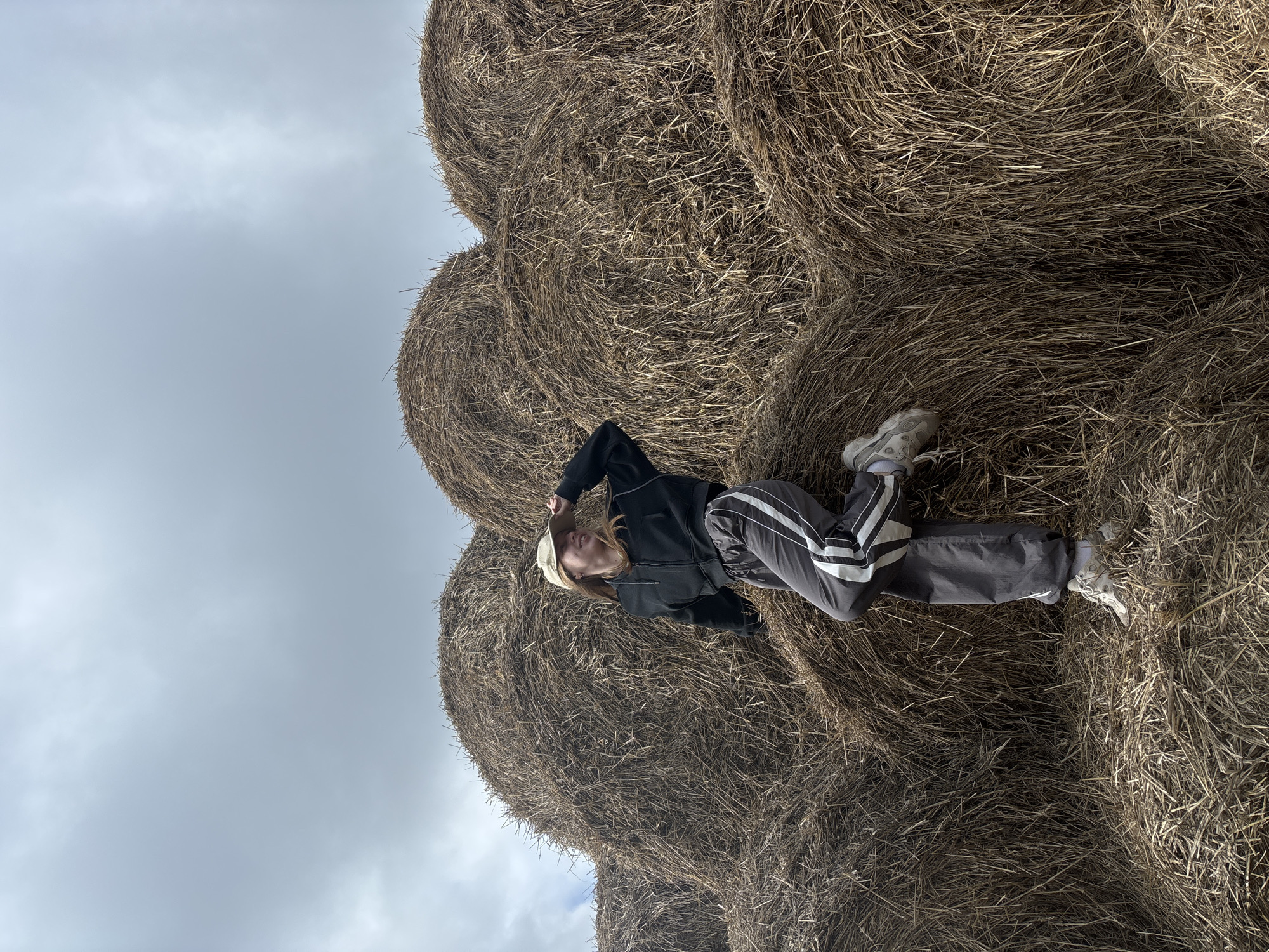 A person is lying on top of a stack of hay bales. The individual is wearing a dark jacket, gray pants with white stripes, white sneakers, and a light-colored baseball cap. The sky is overcast. The image was taken outside the city of Mokhnutino, Russia, as implied from the description.